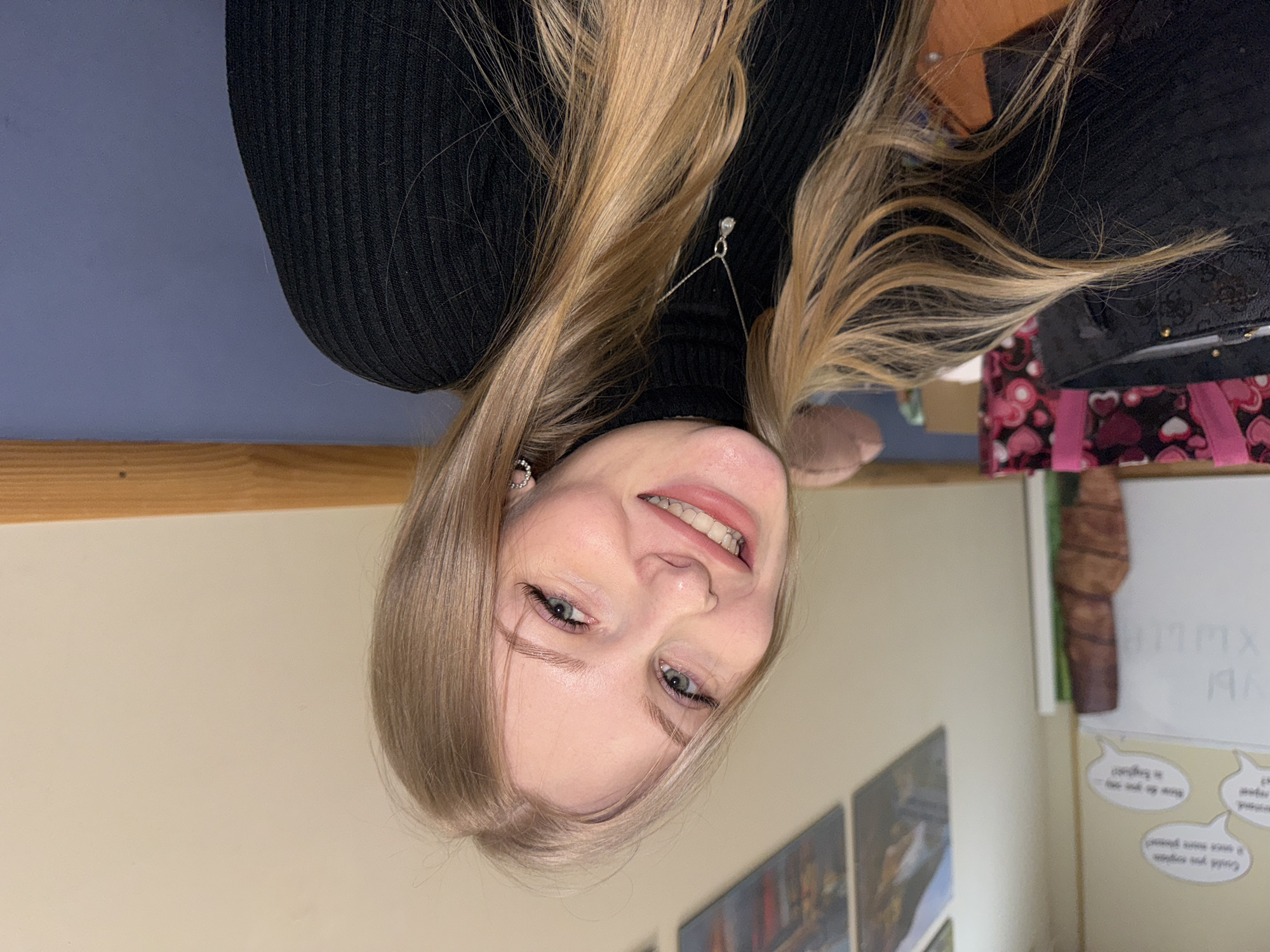 The image, presented upside down, features a young woman smiling and looking directly at the camera. She has long, straight blonde hair, blue eyes, and fair skin, with light makeup accentuating her features. She is wearing a black ribbed turtleneck top. A delicate silver chain with a small clear pendant hangs around her neck, and a small hoop earring with rhinestones is visible in her left ear. Her expression is joyful and relaxed.
The setting is indoors, likely a classroom or an informal office environment. The background consists of walls in light beige/off-white and a muted blue-purple hue, separated by a wooden trim or shelf. To the right of the woman, a white board is partially visible with faint, upside-down markings, including "XMMX". Below the whiteboard, several speech bubble cutouts are affixed to the wall, containing partially legible English text such as "Could you..." and "Can you say...". Other background elements include a patterned fabric with pink and white hearts, a dark object resembling a bag, a decorative wooden item, and partially visible framed pictures on the wall. The overall scene suggests a casual indoor moment in Perm, Russia.