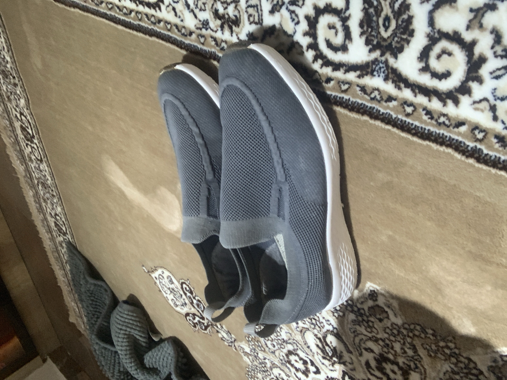 A pair of dark gray slip-on athletic shoes are placed on a beige rug with an ornate, traditional pattern. The shoes are positioned diagonally across the frame, with the front of the shoes pointing towards the bottom right. The rug features a central field of light brown and darker brown, beige, and black intricate scrollwork and floral motifs, bordered by a narrower band of similar patterns. To the left of the shoes, a crumpled, textured, dark gray blanket or fabric is visible, partially obscuring the rug. The lighting appears to be dim, suggesting an indoor setting. The image does not provide any visual cues to indicate the specific location within Abuja, Nigeria.