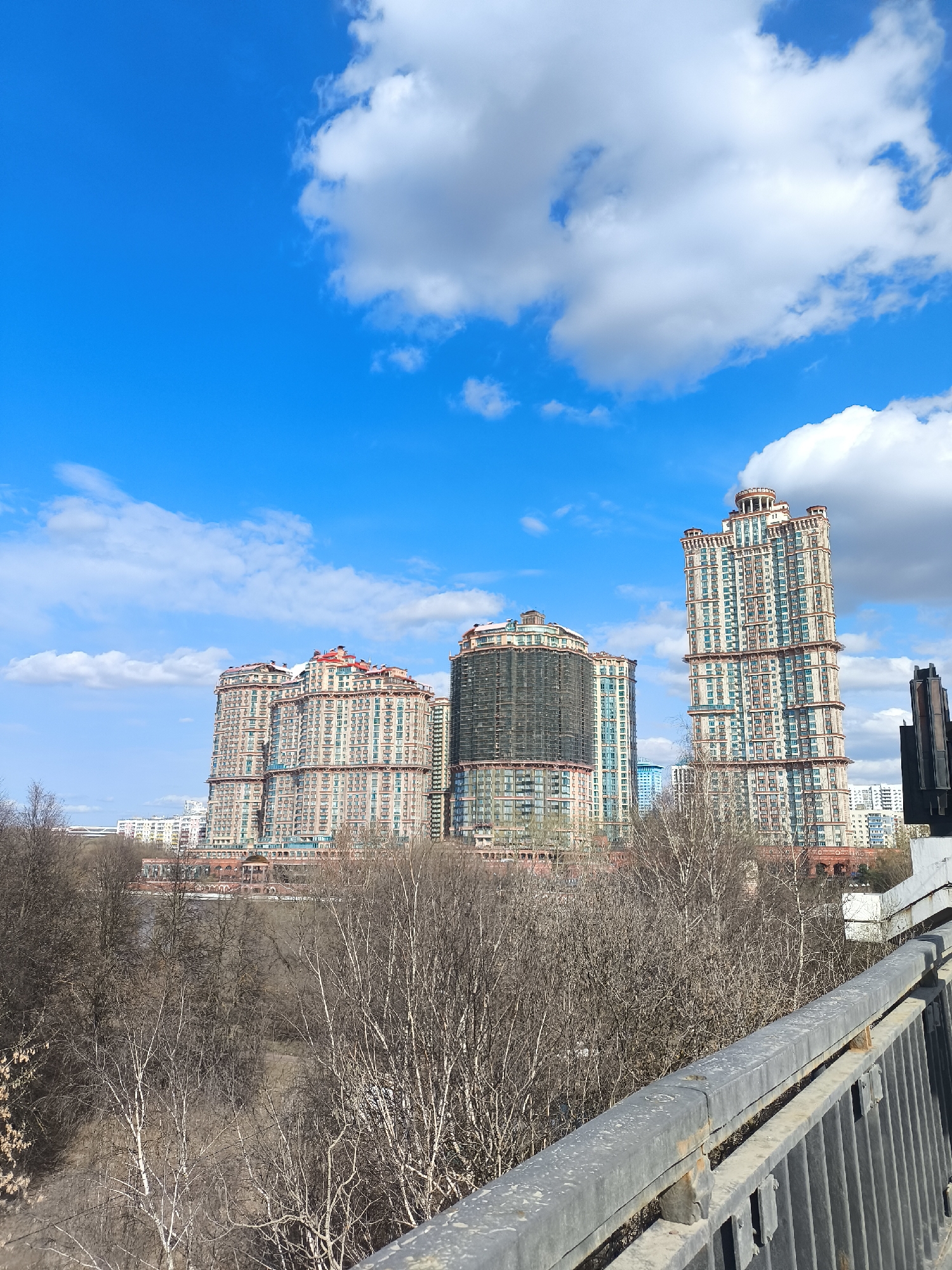 The image captures a view of several high-rise buildings in Khimki, Russia, under a bright blue sky with scattered white clouds. The buildings have a patterned facade with terracotta and beige tones and are distinct in height and design, with the tallest one on the right having a distinctive crown-like top. A partially covered building is in the center of the group. In the foreground, there are bare trees, indicating it might be late autumn or early spring. A metal railing with signs of rust stretches across the bottom of the frame, suggesting the photo was taken from a bridge or an elevated position. The overall scene is urban, with a mix of natural and built environments.
