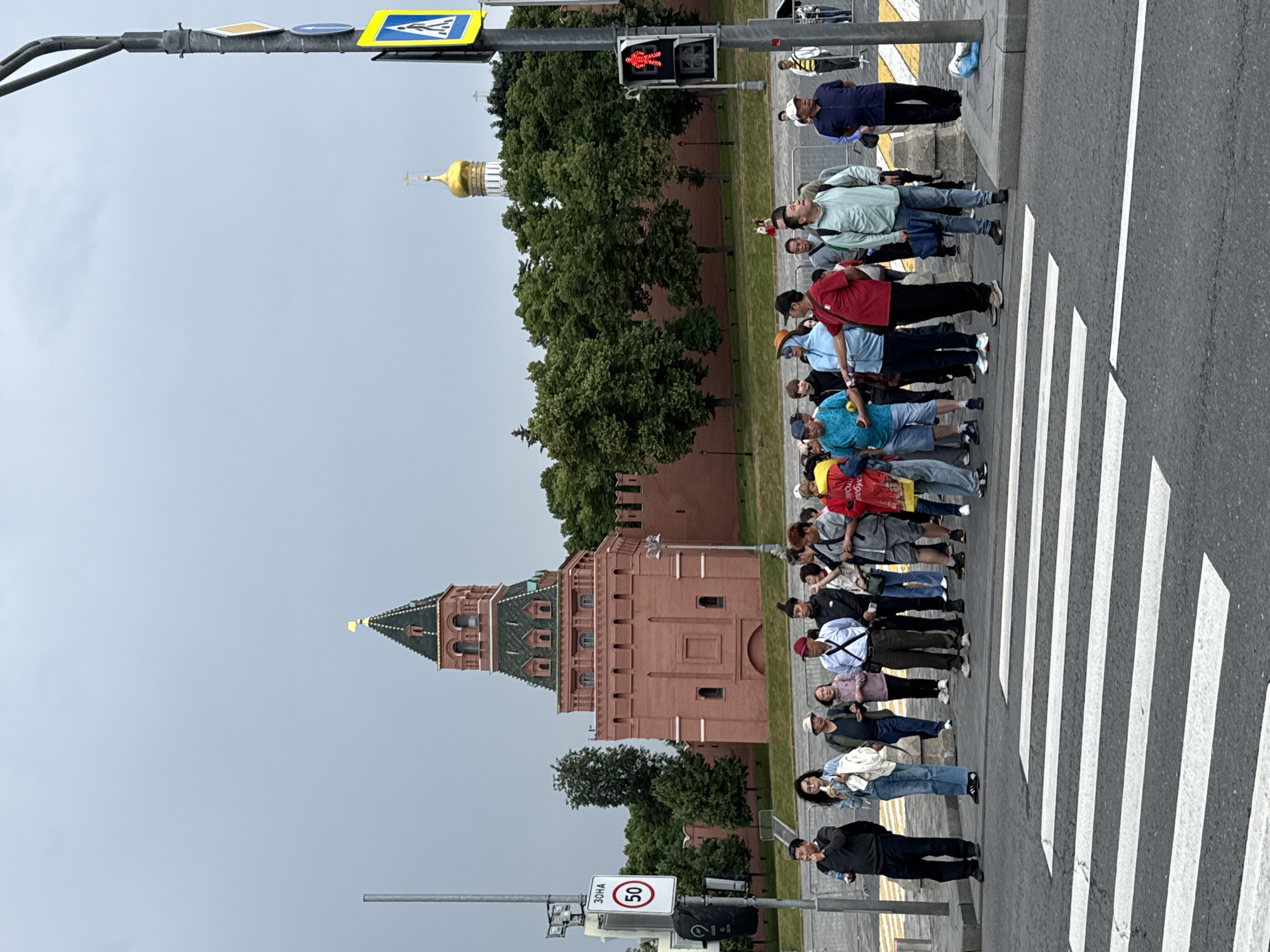 In Moscow, Russia, on a cloudy day, a group of tourists stands at a pedestrian crossing. Behind them, the iconic red brick walls of the Kremlin and a red tower with a green roof and a golden spire are visible. To the right of the tower, a golden-domed church rises above lush green trees. A traffic light on a pole displays a red pedestrian signal. People of various ages and ethnicities are dressed in casual clothing, with some holding bags or looking around. The crossing is marked with white stripes on asphalt. A speed limit sign indicating "50" is posted on a pole with a street sign reading "ЗОНА."