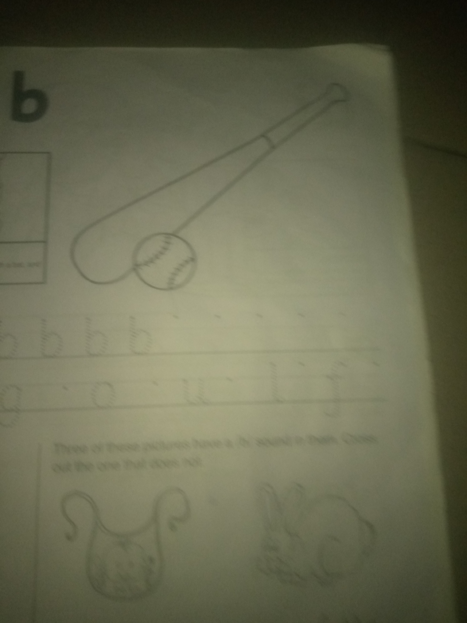 The image displays a page from a children's educational worksheet. Prominently featured is the letter "b" in a large, bold font at the top left. To the right of the letter "b" is a line drawing of a baseball bat and a baseball. Below this are lines of dotted letters, likely for tracing practice, showing lowercase "b"s. Further down, there are more dotted letters, and then a sentence is written: "Three of these pictures have a /b/ sound in them. Cross out the one that does not." Below this sentence are two simple line drawings of objects. The first is a bib, and the second is a rabbit. The image is a photograph of a paper document and is not located in Jauro Yinu, Nigeria.
