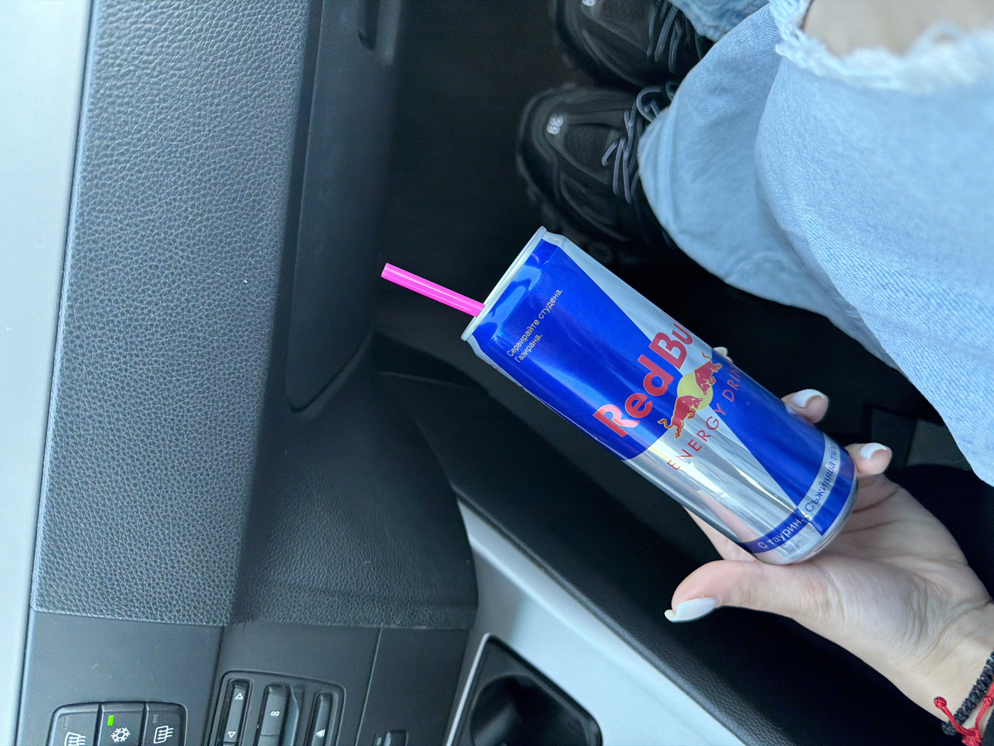 A right-handed person with pale skin and white manicured nails is holding a can of Red Bull with a pink straw in the interior of a car. The can of Red Bull is blue and silver with the words "Red Bull ENERGY DRINK" printed on it. The text on the can is in Bulgarian, including the phrases "Сервирайте студена. Газирана" which translates to "Serve cold. Carbonated" and "С таурин. Съживява тяло" which translates to "With taurine. Revitalizes the body." The hand is adorned with a black and red bracelet. Above the Red Bull, the person is wearing faded blue jeans and black and grey sneakers. The car interior is dark with grey and black textured panels. The location is assumed to be in Letnitsa, Bulgaria based on the Bulgarian text on the can.