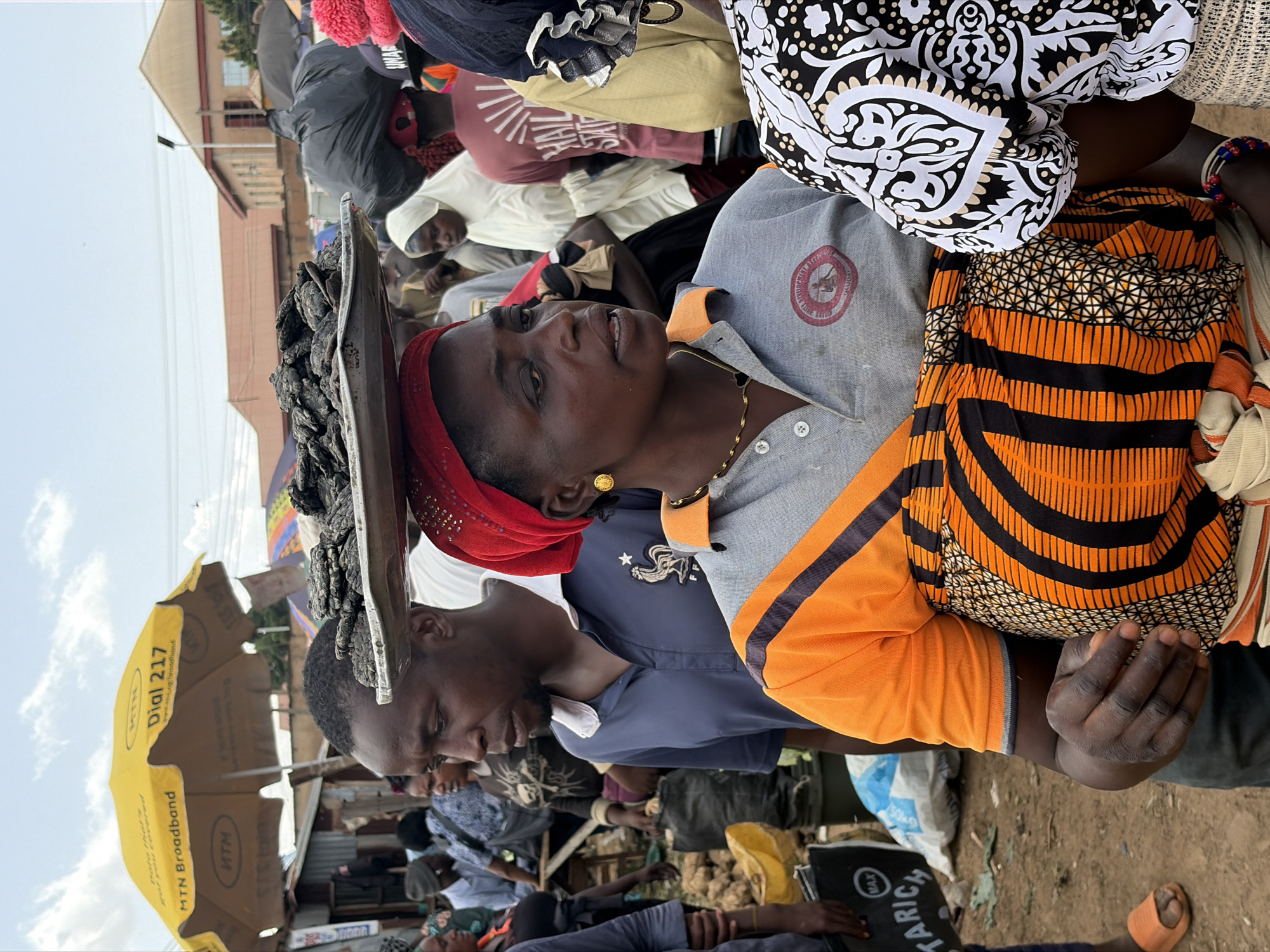 In Gura Top, Nigeria, a group of people are gathered in an outdoor market under a partly cloudy sky. The central figure, a woman with a red headscarf, is wearing a grey polo shirt with an orange and black patterned wrap around her waist. She is looking to her right, with her mouth slightly open as if speaking. In front of her, a man wearing a blue polo shirt carries a tray of dark, lumpy items on his head. Behind the central woman, other market-goers are visible, some carrying large bundles and others wearing headscarves. A yellow umbrella with the MTN logo is prominent in the background. The ground appears to be dirt and scattered debris, with various stalls and structures suggesting a busy marketplace environment.