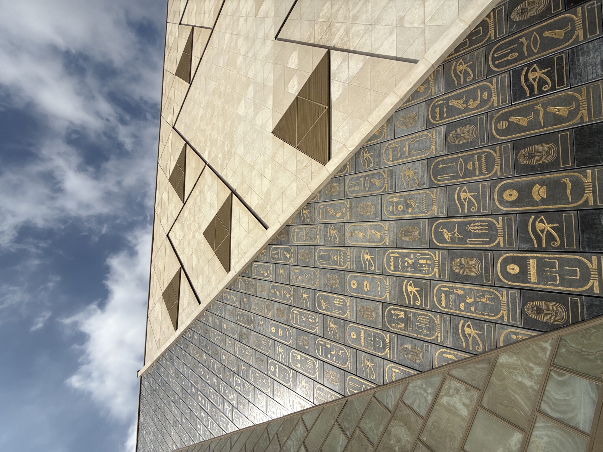 This is a low-angle shot of the exterior of the Grand Egyptian Museum in Kafrat Nassar, Egypt. The building's facade is a striking architectural design featuring a combination of materials and geometric patterns. A significant portion of the image is dominated by a section of the building covered in dark rectangular panels, each inscribed with golden hieroglyphic symbols. These panels are arranged in vertical rows, creating a visually rich texture.
Adjacent to this section, another part of the facade is clad in light-colored, square tiles arranged in a diamond pattern. Interspersed within this tiled area are triangular bronze accents, adding another layer of geometric complexity and visual interest.
In the upper left portion of the frame, a glimpse of the sky is visible, showing a mix of blue sky and scattered white clouds. The overall impression is one of modern architecture drawing inspiration from ancient Egyptian motifs, set against a backdrop of a partly cloudy sky. There are no people visible in the image.