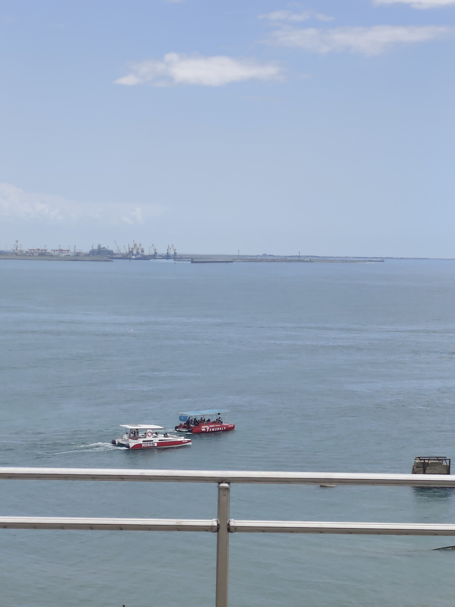 The image shows a wide expanse of blue sea under a light blue sky with a few scattered white clouds. In the middle ground, two small boats are cruising on the water. The boat on the left is a white catamaran with red accents and the word "MONACO" visible on its side. It appears to be moving at a moderate speed, leaving a small wake. The boat on the right is red and has a canopy providing shade for passengers. The word "TAMSHALY" is visible on its side. Several people are seated in this boat.
In the background, a distant industrial port is visible on the horizon, characterized by numerous cranes and structures. The water is calm, suggesting a pleasant day. The light suggests it is daytime, likely late morning or early afternoon. The overall scene depicts a leisurely day at sea near a port area. The presence of the boats and the industrial port provides context of a coastal location with both recreational and industrial activity.