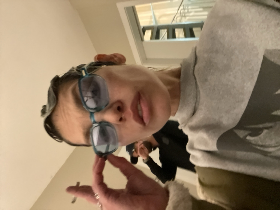 A selfie-style photograph taken indoors in Tel-Aviv, Israel, features two individuals. In the foreground, an adult female looks directly towards the camera. She has dark, short, styled hair and wears teal-framed sunglasses with light blue lenses, which she adjusts with her right hand. Her right hand displays multiple rings, and a lit cigarette is held between her index and middle fingers. She wears a grey turtleneck sweatshirt with a dark graphic print partially visible on the chest, and a green outerwear garment with a light-colored collar is visible on her left shoulder. Her lips are slightly parted. In the blurred mid-ground, an adult male stands behind her, wearing a dark cap and a dark long-sleeved shirt or jacket, with his right hand raised towards his face. The background consists of a light-colored wall, and to the right, an open doorway reveals a bright interior space with a multi-story staircase and a light-colored railing.