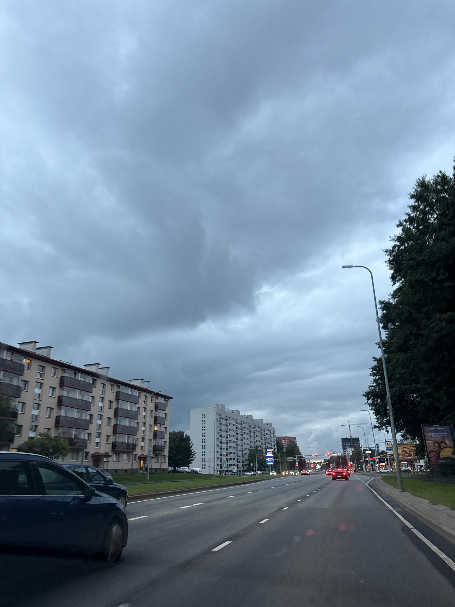 The image depicts a view of a multi-lane road in Tallinn, Estonia, under an overcast sky. The road is lined with several tall apartment buildings on the left side of the frame, featuring balconies and rows of windows. On the right side, there are trees and a lamppost. Traffic is visible on the road, with several cars driving in the same direction. In the distance, traffic lights and more buildings can be seen. A billboard for a fast-food restaurant is positioned on the right side of the road. The overall atmosphere is one of an urban environment on a cloudy day.