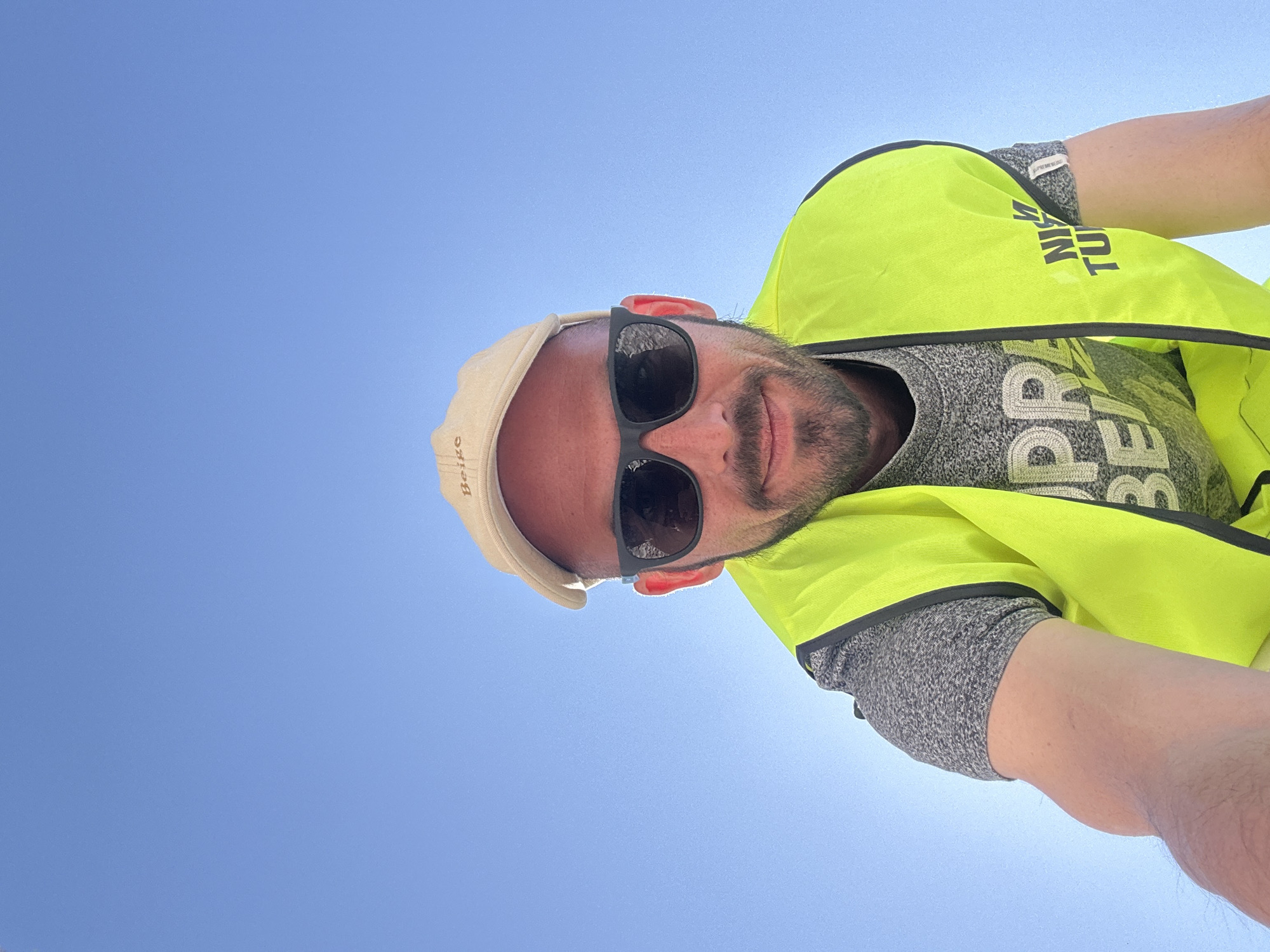 A ground-level selfie shows a man with fair skin, sunglasses, and a light-colored hat. He has a beard and appears to be smiling. He is wearing a yellow safety vest over a gray shirt with the word "PREPARED" partially visible. The background is a clear blue sky. The location is unknown.