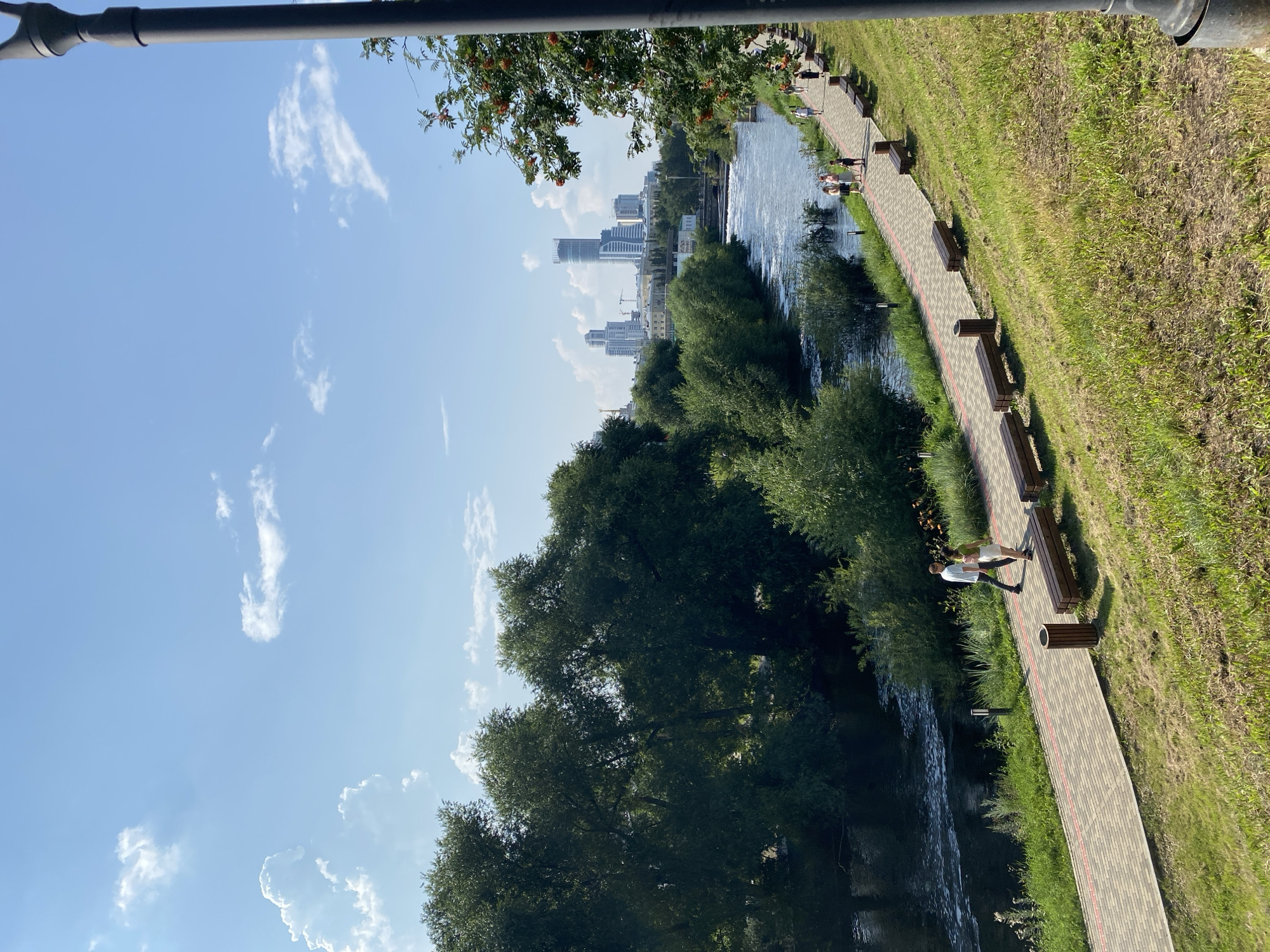 The image depicts a scenic riverside promenade in Yekaterinburg, Russia, likely on the Iset River embankment. In the foreground, a paved walkway curves along a grassy incline. Several wooden benches are strategically placed along the path, inviting rest. A couple is walking hand-in-hand on the walkway, while other individuals are scattered further down, strolling or sitting.
The river flows to the left, bordered by lush green trees and vegetation. The water is calm in some areas, with small ripples and white water indicating a slight current. Across the river, a dense line of trees forms a natural barrier, beyond which the Yekaterinburg city skyline rises. Several modern skyscrapers dominate the view, including a distinctive tall, cylindrical building. The sky is a clear blue with scattered white clouds, suggesting a pleasant, sunny day. A dark lamppost is partially visible on the right edge of the frame, along with branches of a tree with red berries.