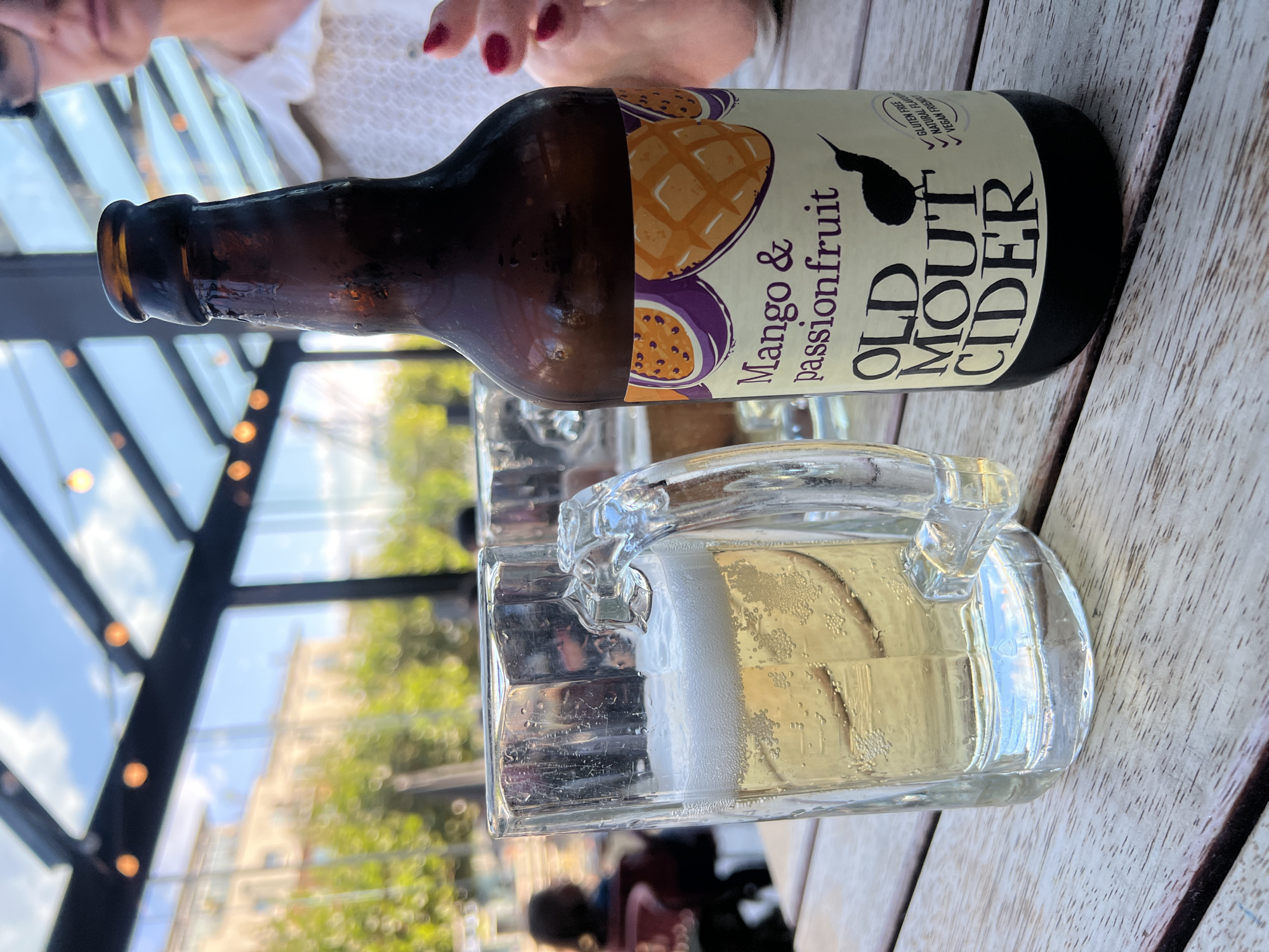 A glass mug of cider sits on a wooden table next to a bottle of Old Mout Cider, Mango & Passionfruit flavor. The glass is partially filled with a pale, bubbly liquid and topped with a frothy head. The bottle, made of brown glass, has a label featuring illustrations of mango and passionfruit, and a black kiwi bird silhouette. In the background, a woman with red nail polish is visible, wearing a white top. The setting appears to be an outdoor seating area with a glass roof, suggesting a restaurant or pub. The scene is bathed in natural light, with trees and buildings visible through the glass. This image was taken in Portsmouth, United Kingdom.