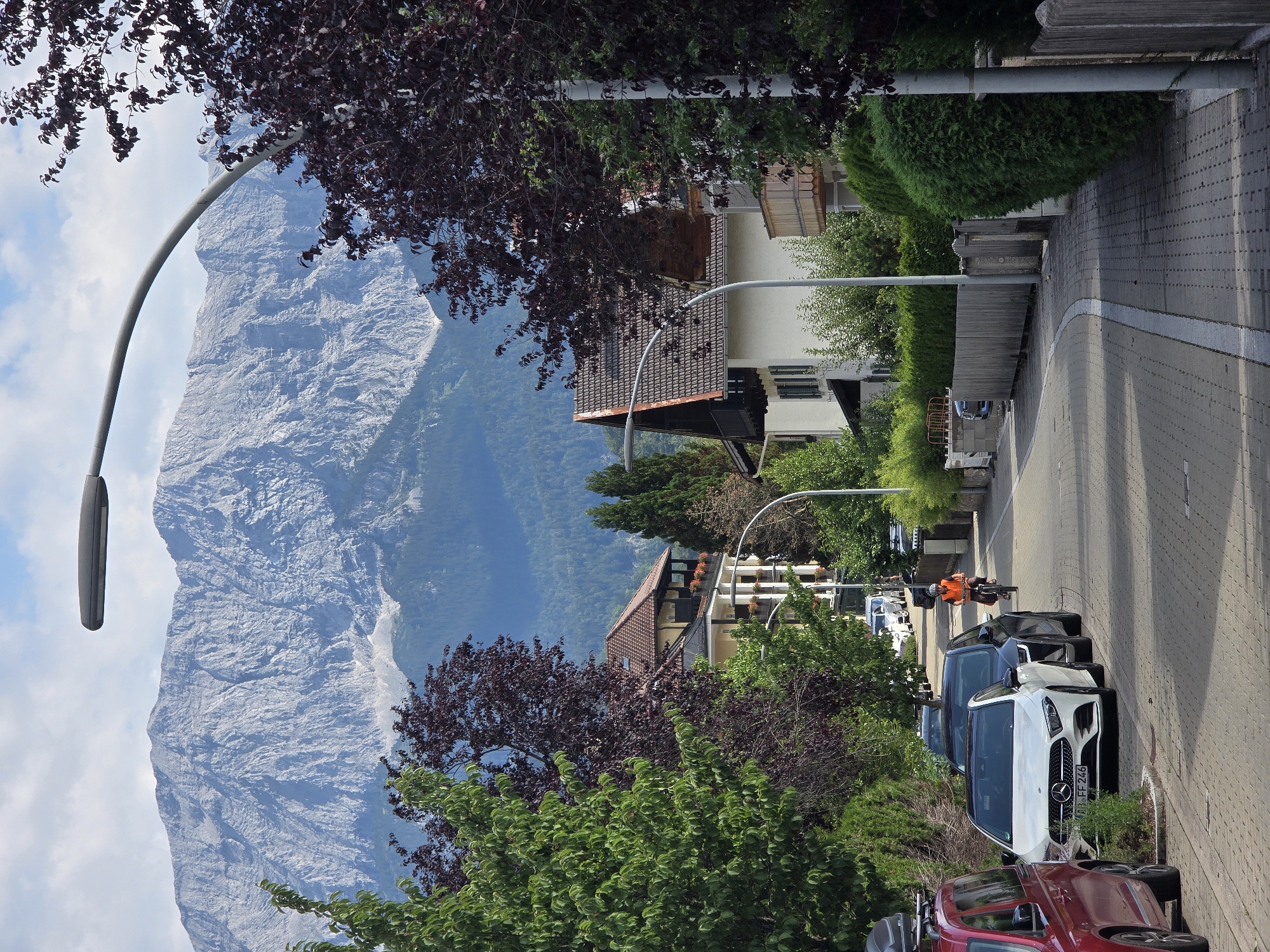The image depicts a street scene in Garmisch-Partenkirchen, Germany, with a prominent mountain range in the background. The foreground features a paved road lined with parked cars. A white Mercedes-Benz, license plate AB FF 246, is parked on the right side of the road. To its left, a dark-colored sedan is parked. Further down the street, a person on a bicycle, wearing an orange vest, is moving away from the camera. Residential buildings with balconies and tiled roofs are visible on the left and right sides of the street, interspersed with lush green trees and hedges. A modern street lamp curves over the road. The sky is partly cloudy. The mountain range, appearing rugged and imposing, dominates the upper portion of the image, with some visible snow patches on its higher slopes. The overall atmosphere is that of a tranquil, suburban environment with a breathtaking natural backdrop.