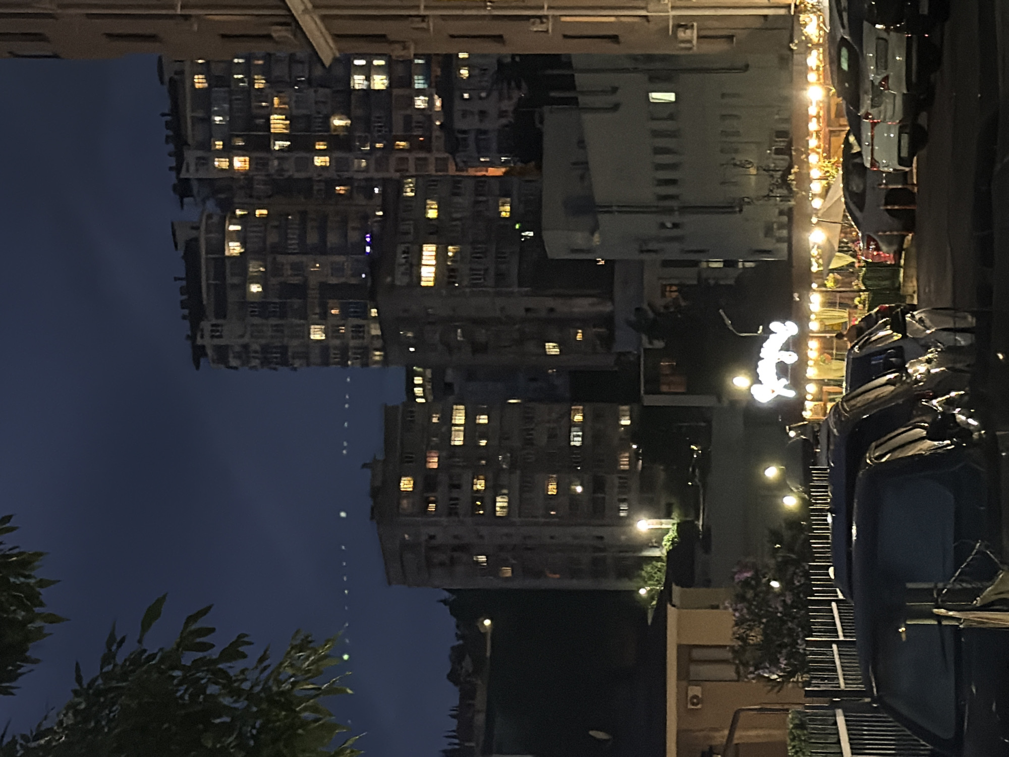 The image depicts an urban nighttime scene in Sochi, Russia, characterized by residential buildings and a street-level establishment. Two prominent apartment complexes with illuminated windows dominate the background. To the left, a lower-rise building with a dark roof and an air conditioning unit is visible. In the foreground, several parked cars are present, with the rear of a dark-colored vehicle partially obstructing the view. A street-level restaurant or cafe, illuminated by strings of lights, is situated behind a black metal fence. The sky is dark, suggesting it is late evening or night.