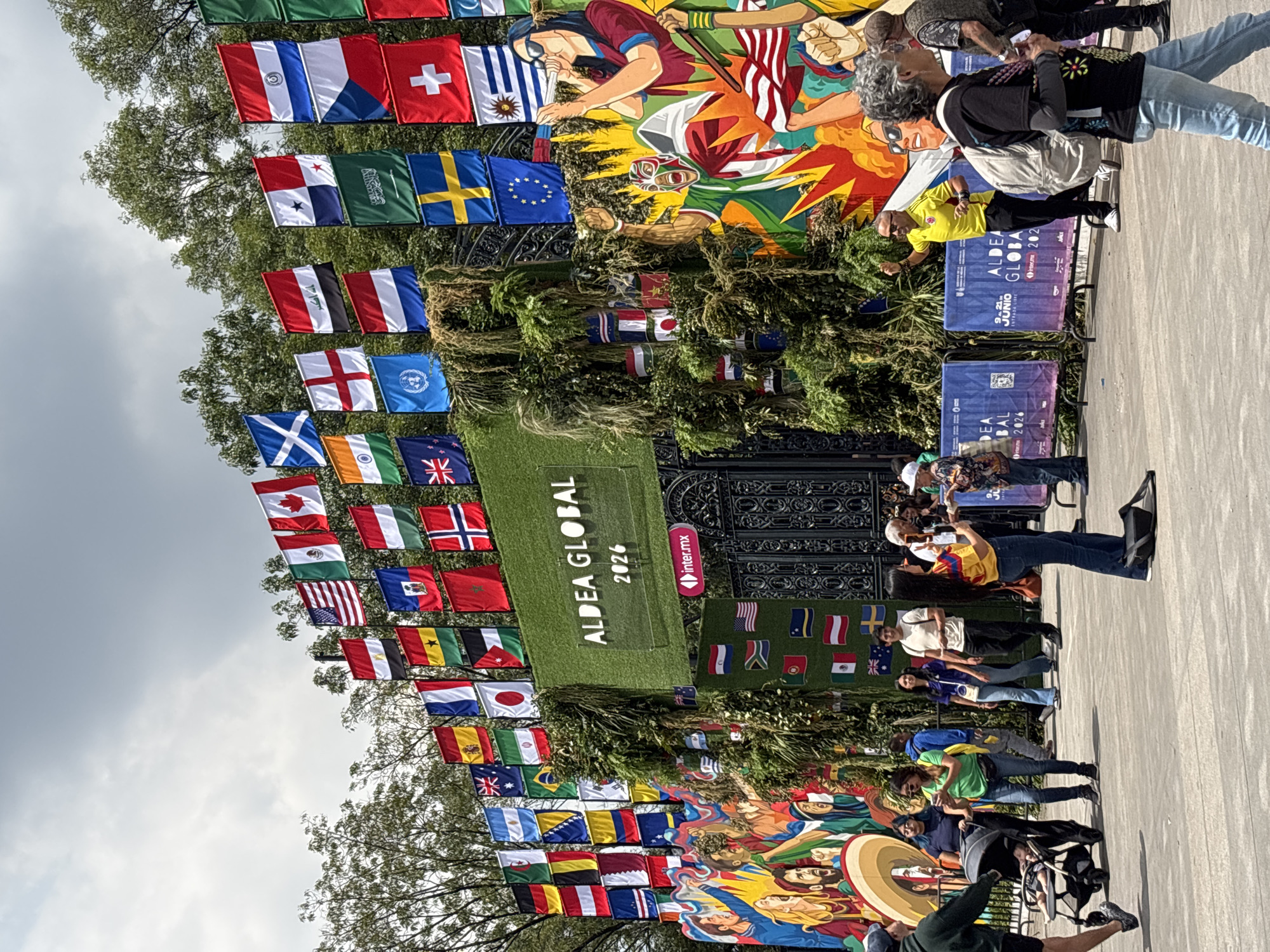 In Mexico City, Mexico, a vibrant event is underway, characterized by a large, ornate entrance adorned with numerous national flags from around the world. The flags, displayed in rows and columns, create a festive and inclusive atmosphere, symbolizing global unity. Above the entrance, a prominent sign reads "ALDEA GLOBAL 2026" in large, illuminated letters, indicating the name and year of the event. The entrance is flanked by lush greenery, adding a natural element to the urban setting.
A diverse crowd of people is present, engaged in various activities. Some are strolling through the area, while others are observing the displays. A family with a baby in a stroller is visible, as is a man in a bright yellow shirt interacting with the crowd. Artistic murals featuring stylized figures and explosive imagery contribute to the dynamic and energetic feel of the event. The scene suggests a lively public gathering, possibly a festival, exhibition, or sporting event, celebrating international diversity and the year 2026. The presence of "inter.mx" on a smaller sign suggests sponsorship or partnership by a Mexican telecommunications company. The sky is overcast, indicating a cloudy day.