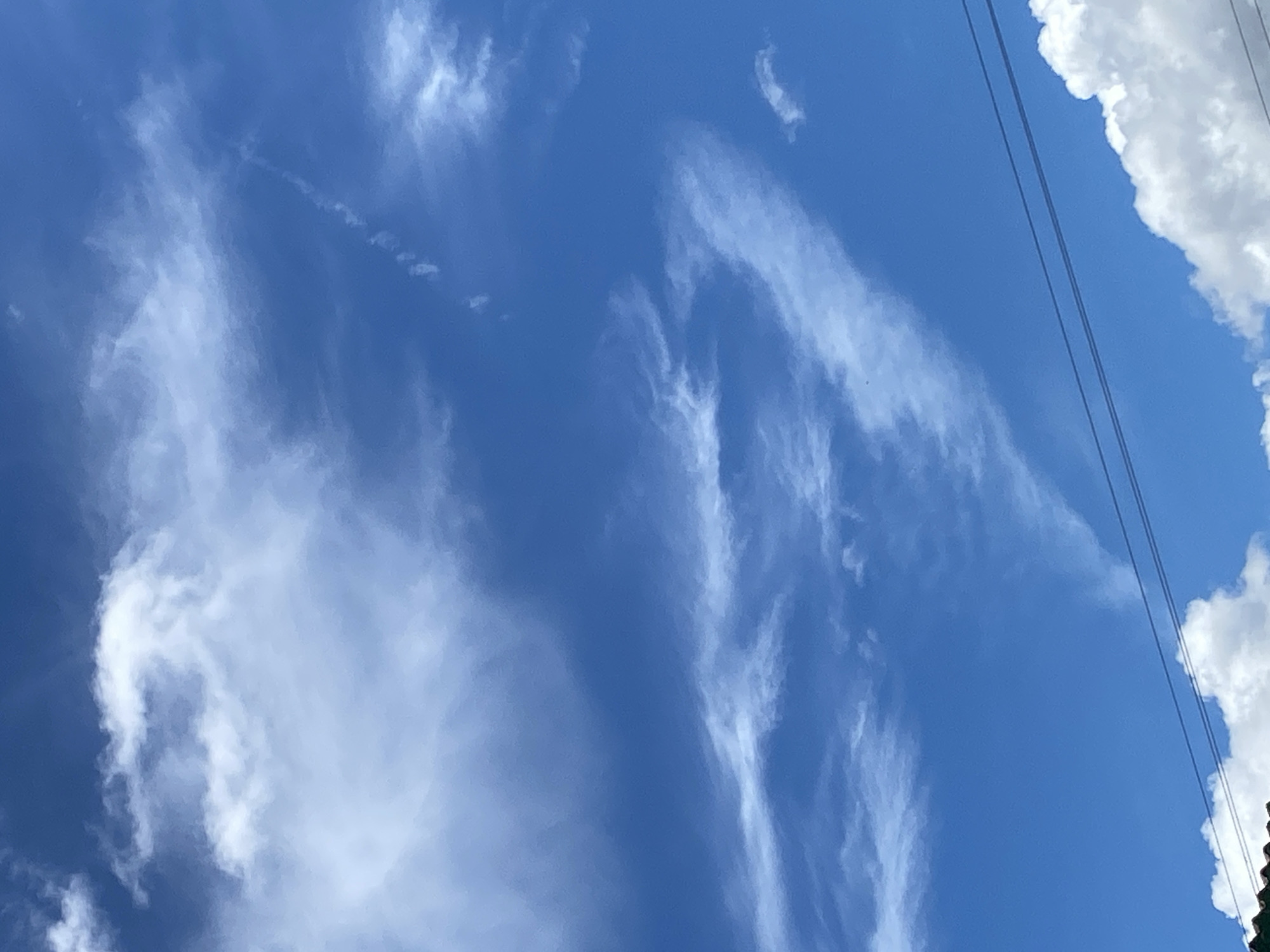 The image is a photograph of a clear blue sky with scattered white clouds. In the lower portion of the image, there are two parallel dark lines that appear to be power lines, and the edge of a roof is visible on the far left. The overall scene suggests an outdoor environment, possibly in a city given the presence of the power lines and roof, though no specific landmarks or identifying features of Tinaquillo, Venezuela are visible in this particular frame. The clouds are wispy and appear to be high in the atmosphere, suggesting fair weather conditions. There are no people or discernible actions taking place.