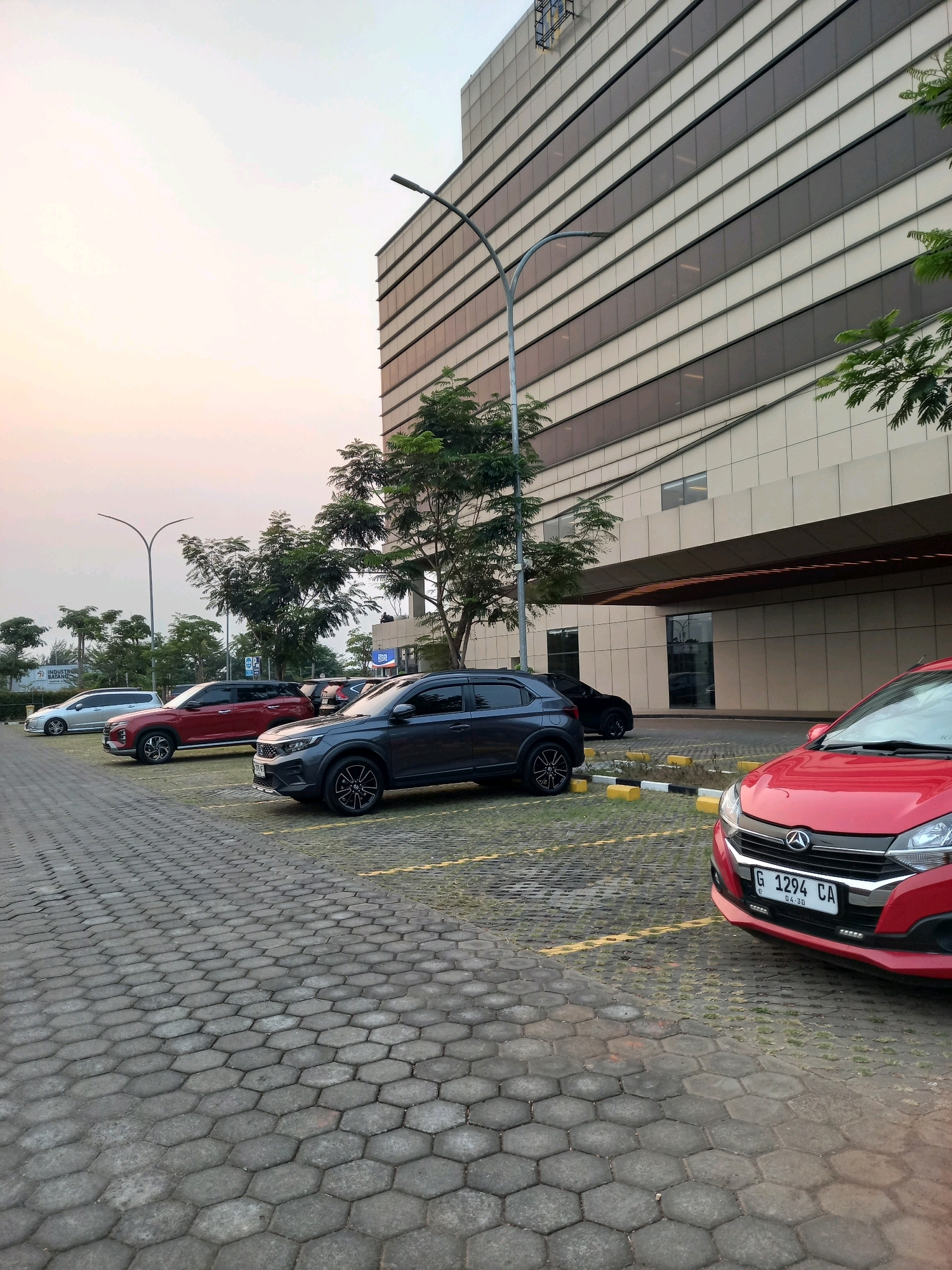 In Ketanggan, Indonesia, this photograph captures a parking lot in front of a modern building. The building features a facade of horizontal beige and dark tinted glass panels. A large street lamp with a curved arm stands to the left of the building. The parking lot is paved with hexagonal grey interlocking concrete blocks and marked with yellow lines. Several cars are parked in the lot, including a red hatchback in the foreground, a dark gray SUV, a red SUV, and a silver sedan further back. Trees with dense green foliage are planted around the parking lot and the building. The sky is a soft gradient of pale orange and white, indicating either sunrise or sunset.