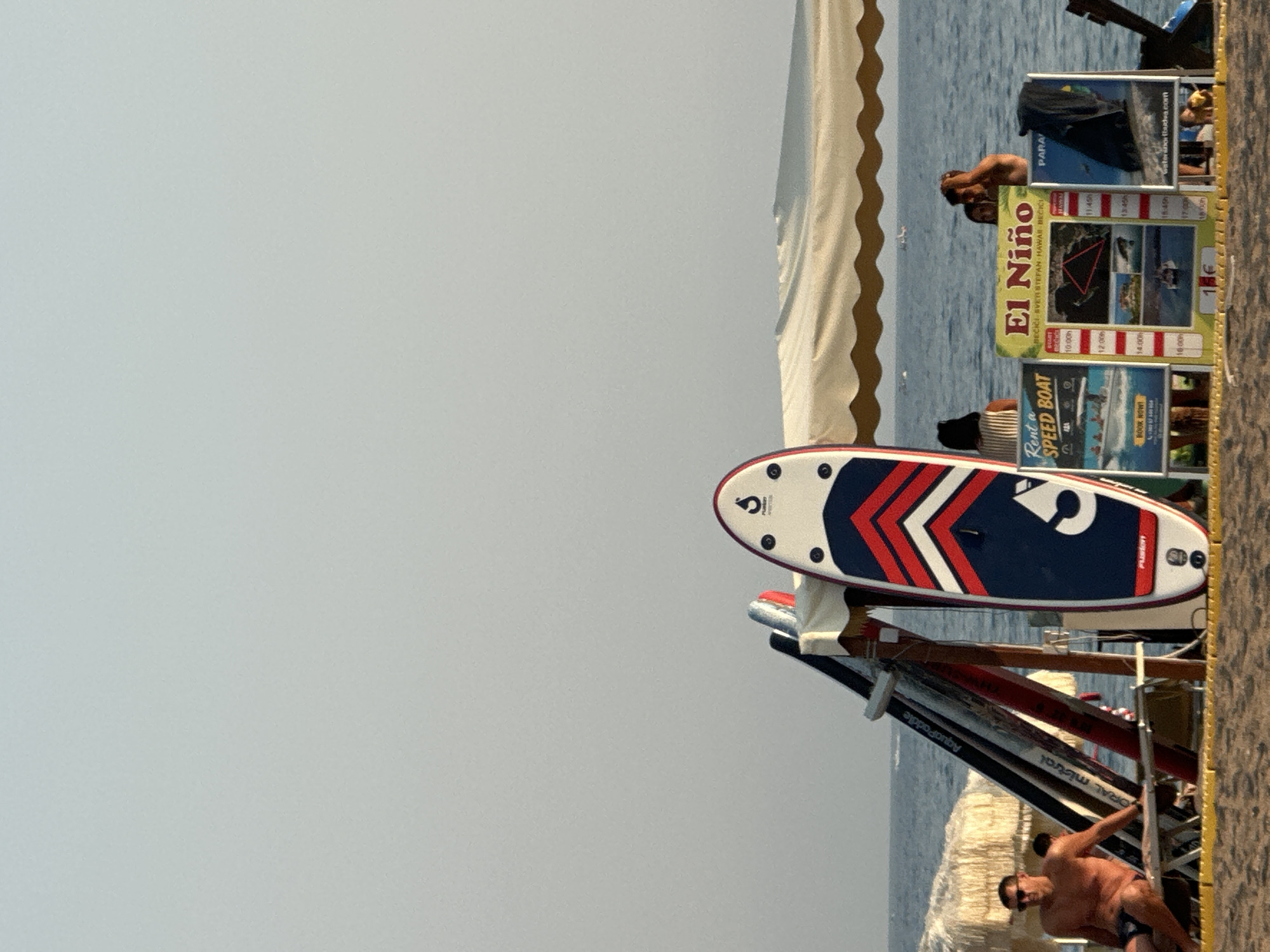 The image depicts a beach scene in Boreti, Montenegro, on a clear day with the sky and sea in the background. In the foreground, several stand-up paddleboards are arranged vertically. One paddleboard, in the center, is white with red and blue chevron designs. To the left, a man wearing sunglasses and swim trunks is seated on a lounge chair. Behind him, other paddleboards and beach umbrellas are visible. In the middle ground, a series of posters are displayed. One poster is yellow and green, titled "El Niño," with a schedule of activities. Another poster to its left advertises "Rent a Speed Boat." To the right of the "El Niño" poster, another poster is partially visible. A couple is standing behind the posters, near the water's edge. The sea is calm, with a few small boats visible in the distance. The beach is sandy.
