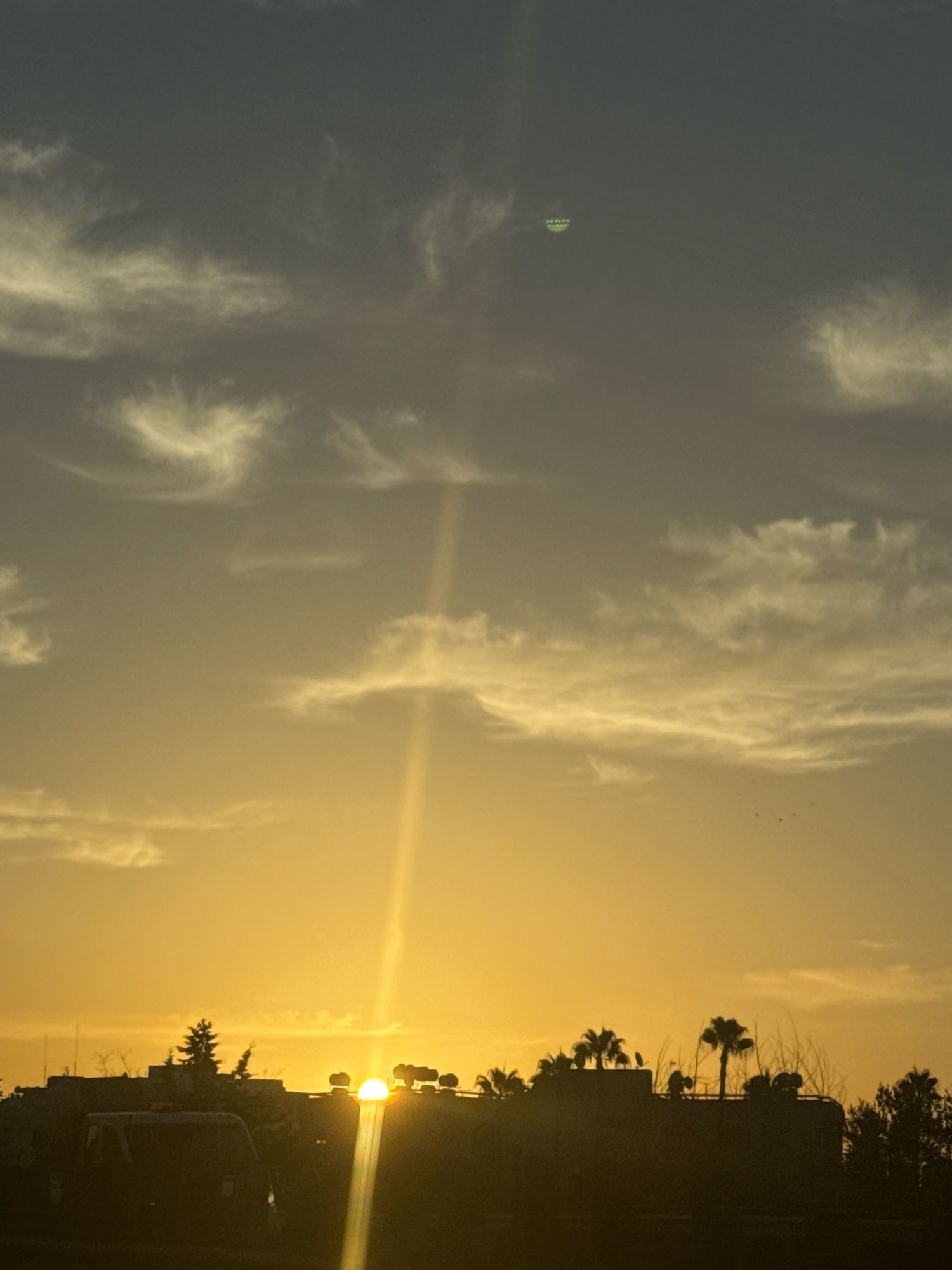 A sunset over Agios Tychona Community, Cyprus, features a radiant sun, partially obscured by silhouetted buildings and trees. A beam of light pierces the sky, illuminating wispy clouds. In the distance, a disc-shaped object with green lights hovers in the sky. A vehicle is visible in the foreground.