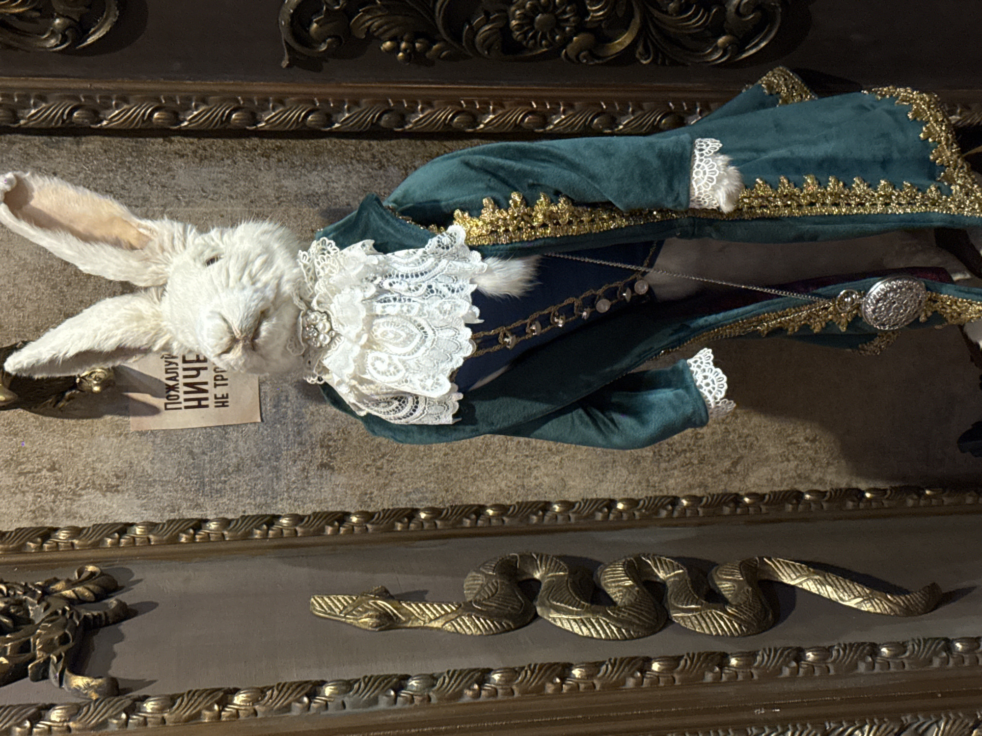 A taxidermied white rabbit, dressed in a green and gold coat with lace trim, lies on its side on a decorative surface. A small rectangular card with Cyrillic script is positioned near the rabbit’s head. The surface features a decorative gold snake and other ornate embellishments. The scene is likely inside a museum or gallery in Moscow, Russia, based on the subject matter and artistic style.