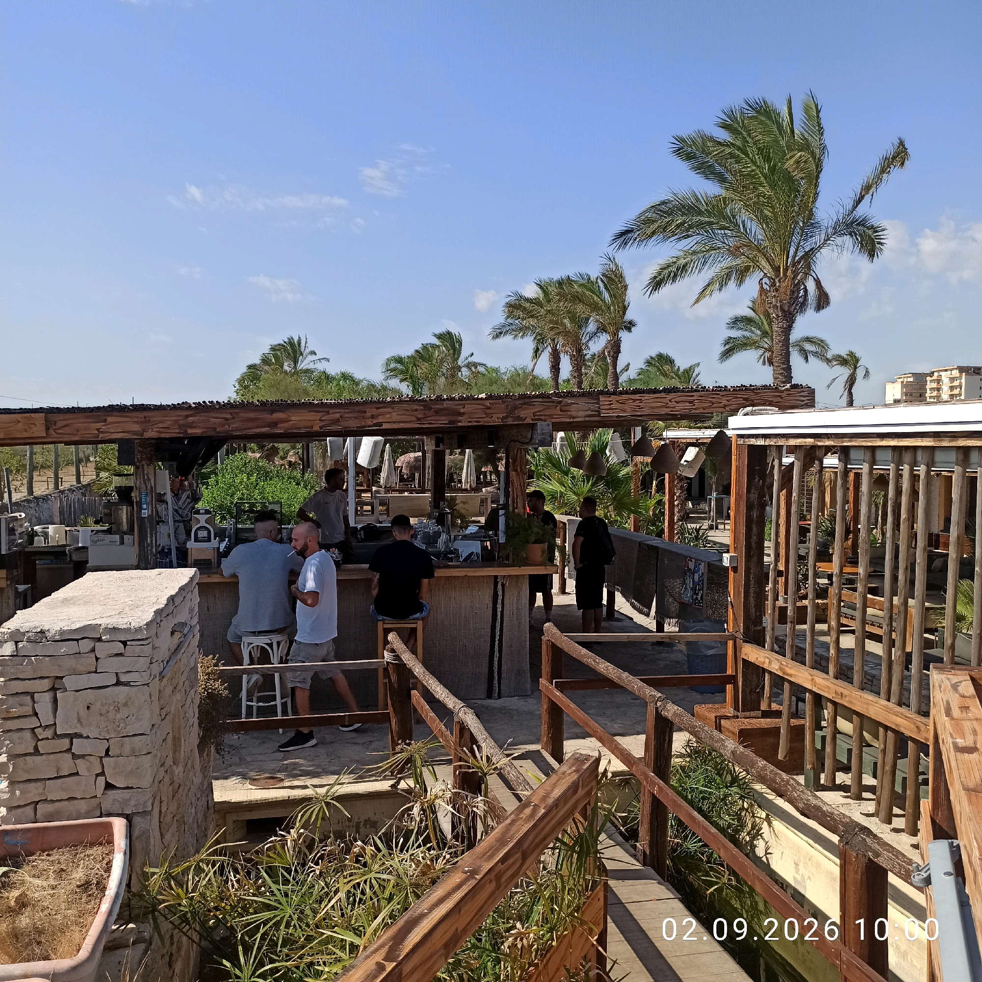 The image depicts an outdoor bar area with a rustic, beach-themed design. Several individuals are present, some standing and some seated at the bar. The bar itself is constructed from wood and natural materials, with a thatched roof overhead. Palm trees and other tropical vegetation are visible throughout the scene, suggesting a warm climate. A wooden walkway with railings leads through the area. In the background, a multi-story building can be seen, indicating the presence of a developed area nearby. The sky is clear and blue, with a few scattered clouds. A timestamp in the bottom right corner reads "02.09.2026 10:00". There are no discernible clues within the image to suggest a specific location in Molfetta, Italy.