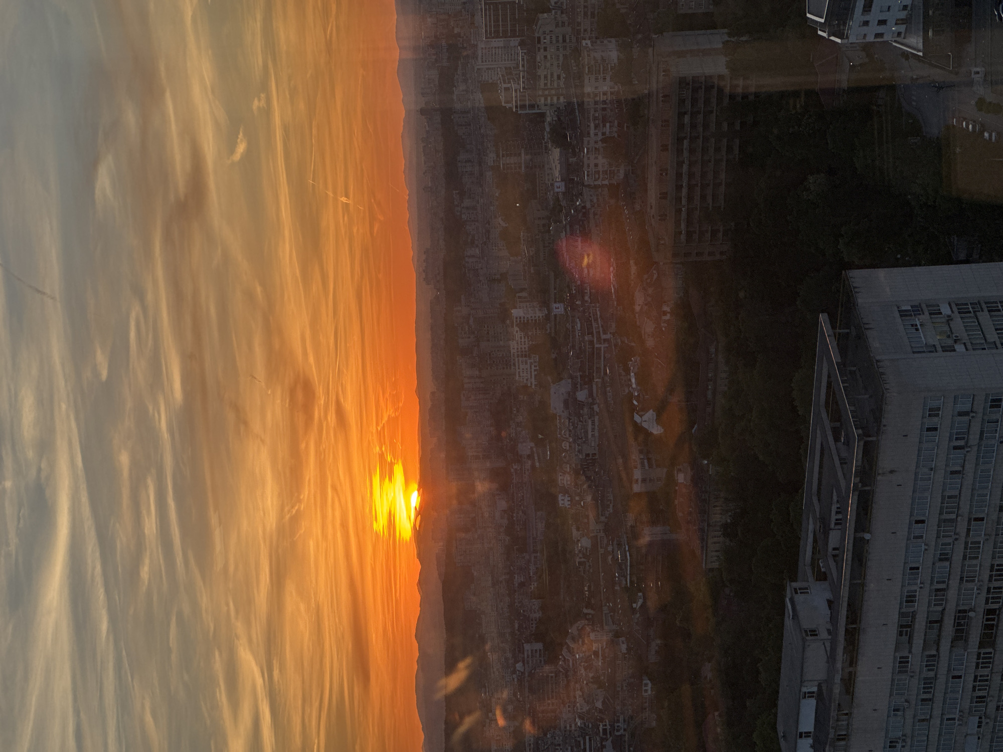 The image depicts a sunset over a city, likely Sofia, Bulgaria, viewed from a high vantage point. The sun is partially obscured by a mountain range on the left, casting an orange glow across the sky and clouds. Below, the city sprawls with a dense array of buildings, including high-rise structures, residential blocks, and commercial areas. A large, modern building is visible in the foreground on the right, suggesting the viewpoint is from another high-rise building. The lighting suggests the photo was taken near sunset, casting shadows and highlighting the building outlines. The sky is a mix of orange, yellow, and grey tones, with striated cloud patterns. There are no visible people or other specific objects, only the general urban landscape. The overall impression is a cityscape at dusk, seen from an elevated perspective.