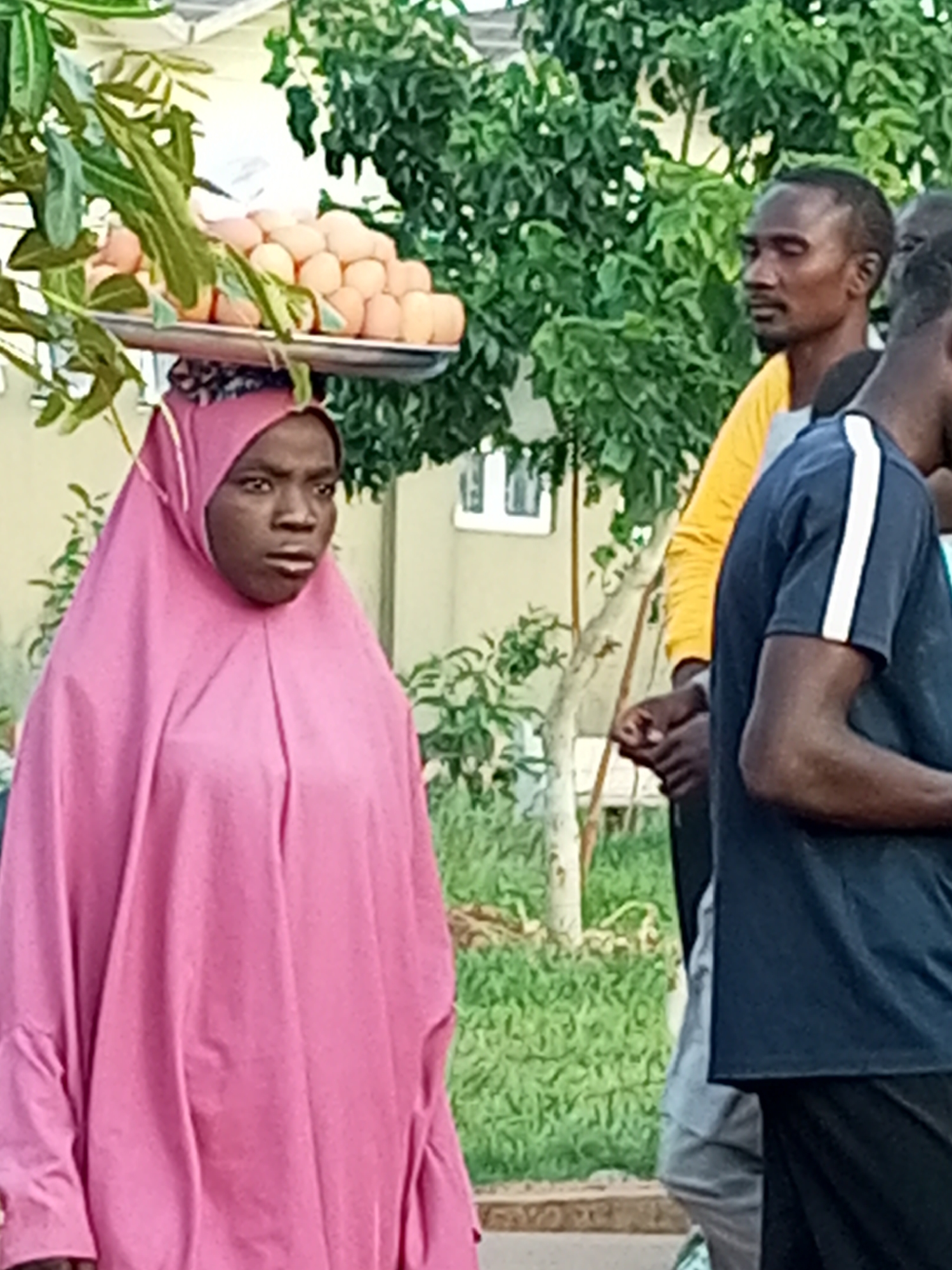 In Gombe, Nigeria, a woman wearing a pink hijab carries a tray of eggs on her head. She is positioned in the foreground, with her face turned slightly to her right. Behind her, a man in a yellow shirt and another person in a dark blue shirt are visible. The background consists of green foliage and a building with windows. The scene appears to be outdoors, possibly on a street or pathway.