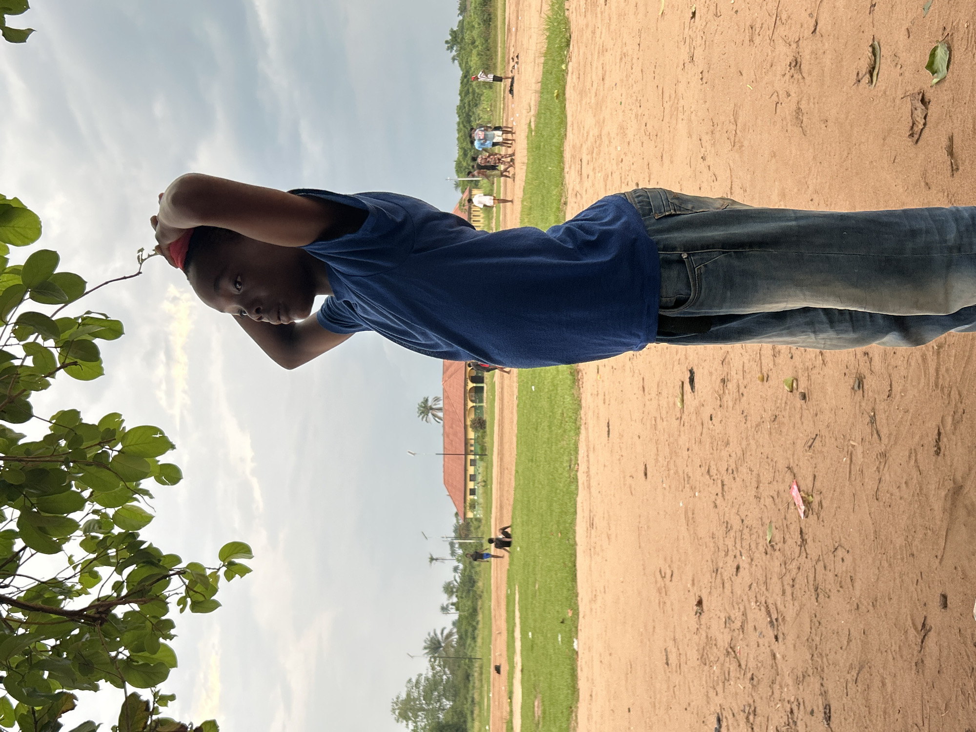 In Ihiagwa, Nigeria, under a cloudy sky and the shade of leafy green branches, a young Black boy stands in profile, facing left, with his hands clasped behind his head. He is wearing a blue t-shirt and blue jeans. The ground in the foreground is sandy and somewhat littered with leaves and debris. In the background, a field with patches of green grass and sandy areas is visible. A building with an orange roof and yellow walls stands in the distance, surrounded by trees and utility poles. Several other individuals, appearing to be children or young adults, are scattered across the field, some standing and others sitting on the grass. One person in the background is sitting on the grass, appearing to be alone, while a group of people are gathered in the middle distance, possibly playing. Another individual is standing on the far right of the field.
