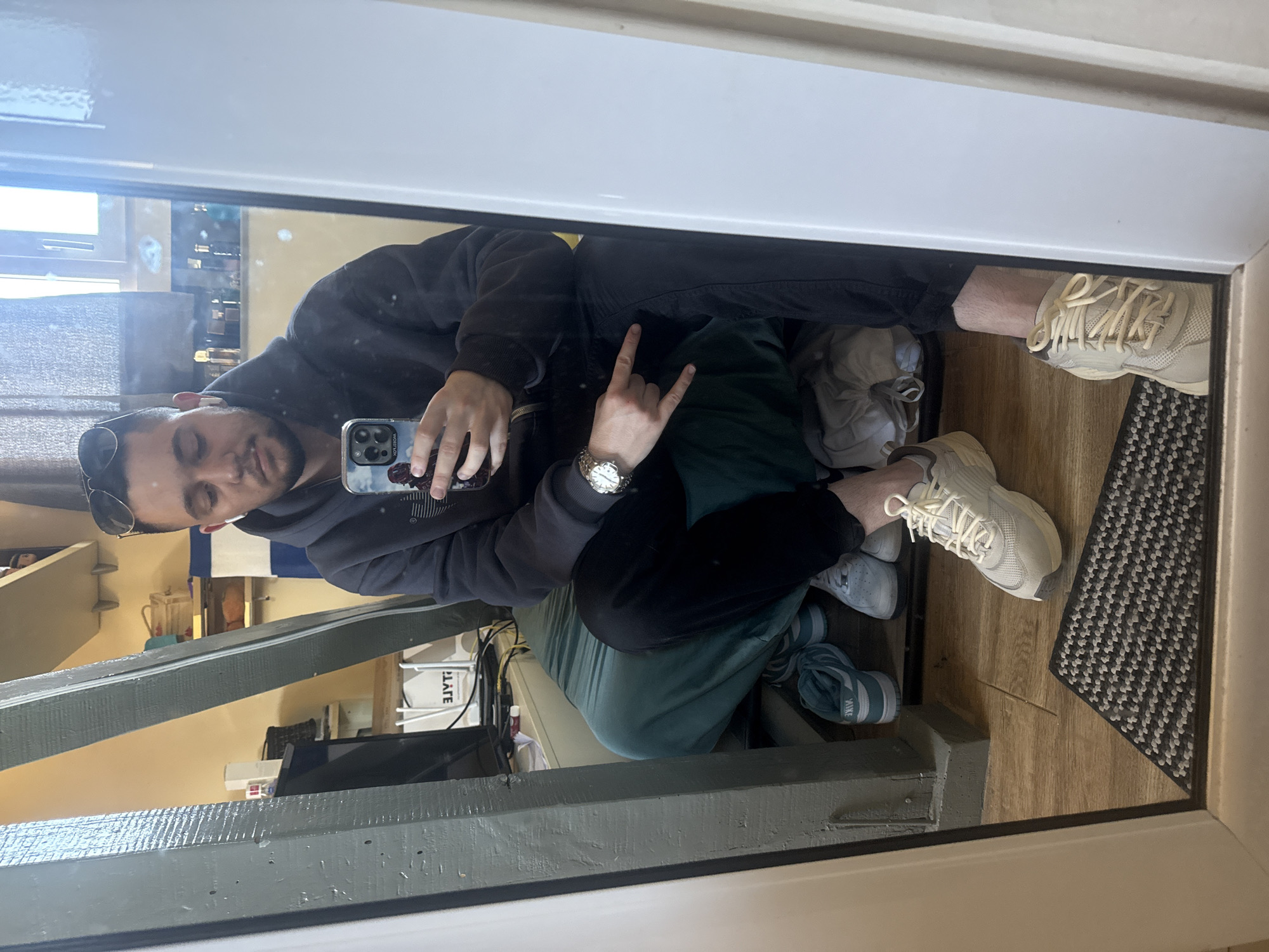 A person takes a selfie in a mirror. The individual, wearing a dark hoodie and pants, holds a phone in one hand and makes a "rock on" gesture with the other. Sunglasses rest on their head. The reflection shows a room with exposed wooden beams, a shelf with various items, and a television. Several pairs of shoes are visible on the floor, along with a patterned rug. The location appears to be indoors. The image does not contain information that identifies the location in the city of Sofia, Bulgaria.