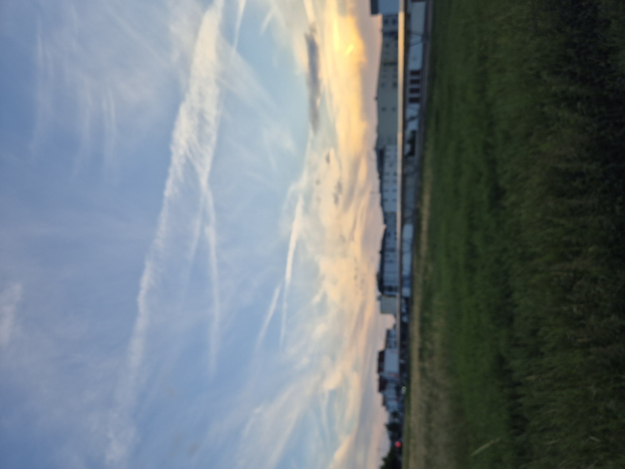 The image depicts a wide, horizontal view of a rural landscape in Ergolding, Germany, at dusk. In the foreground, a dense field of green grass stretches across the frame, showing signs of being mowed. Beyond the field, a long, low-rise building complex is visible, characterized by a light-colored facade and a dark roof. The building appears to be industrial or commercial, with visible doors or entrances. The sky above is a blend of pale blue and soft yellow hues, indicative of sunset. wispy white clouds, possibly contrails, are scattered across the sky, adding texture to the scene. The overall impression is one of tranquility and the transition from day to night in a suburban or semi-rural area. The image is slightly out of focus.
