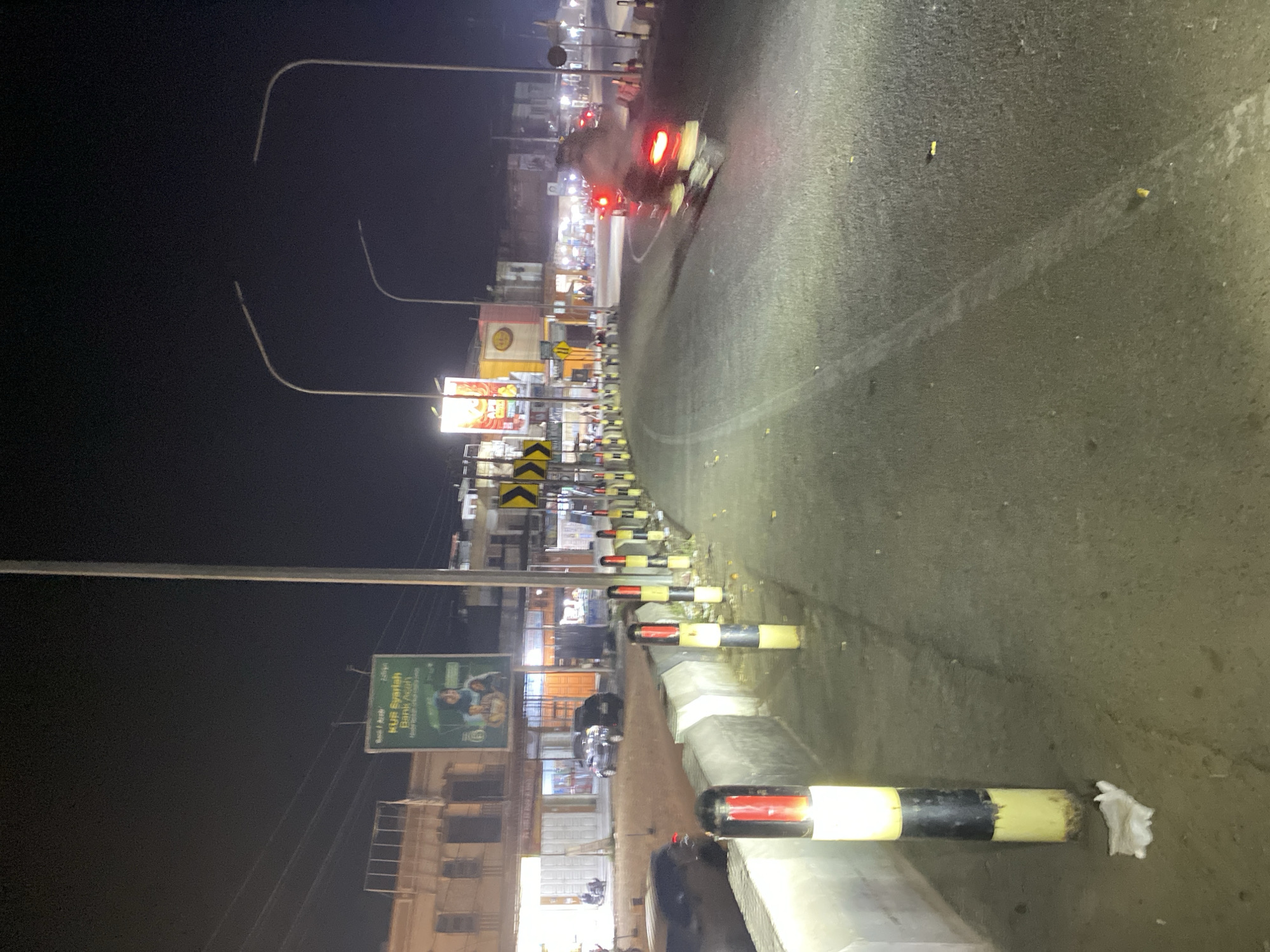 The image depicts a nighttime street scene in an urban or semi-urban environment, likely in Seuneubok Teungoh, Indonesia. The asphalt road, which appears to curve slightly to the right, is illuminated by various light sources.
Along the right side of the road, a series of black and yellow (or red) striped bollards or traffic barriers separate the thoroughfare from a paved curb. Some white debris is visible on the ground near these barriers. On the road itself, a blurred motorcycle with distinct red taillights is captured in motion, indicating ongoing traffic.
The left side of the street features multiple buildings, likely housing shops or businesses, marked by their illuminated windows and signs. Overhead, several streetlights with long, curved arms cast light onto the scene against the dark night sky. Utility poles and overhead power lines are also present. Among the visible signage is a prominent green billboard displaying the text "KUB Syariah Bank Aceh" along with the slogan "Hidup Berkah Dunia Akhirat." Additionally, yellow and black chevron signs are mounted along this side of the road, typically indicating a curve or hazard.
The scene conveys a sense of moderate activity in an urban setting after dark, with artificial lighting providing visibility for vehicular and commercial operations.
