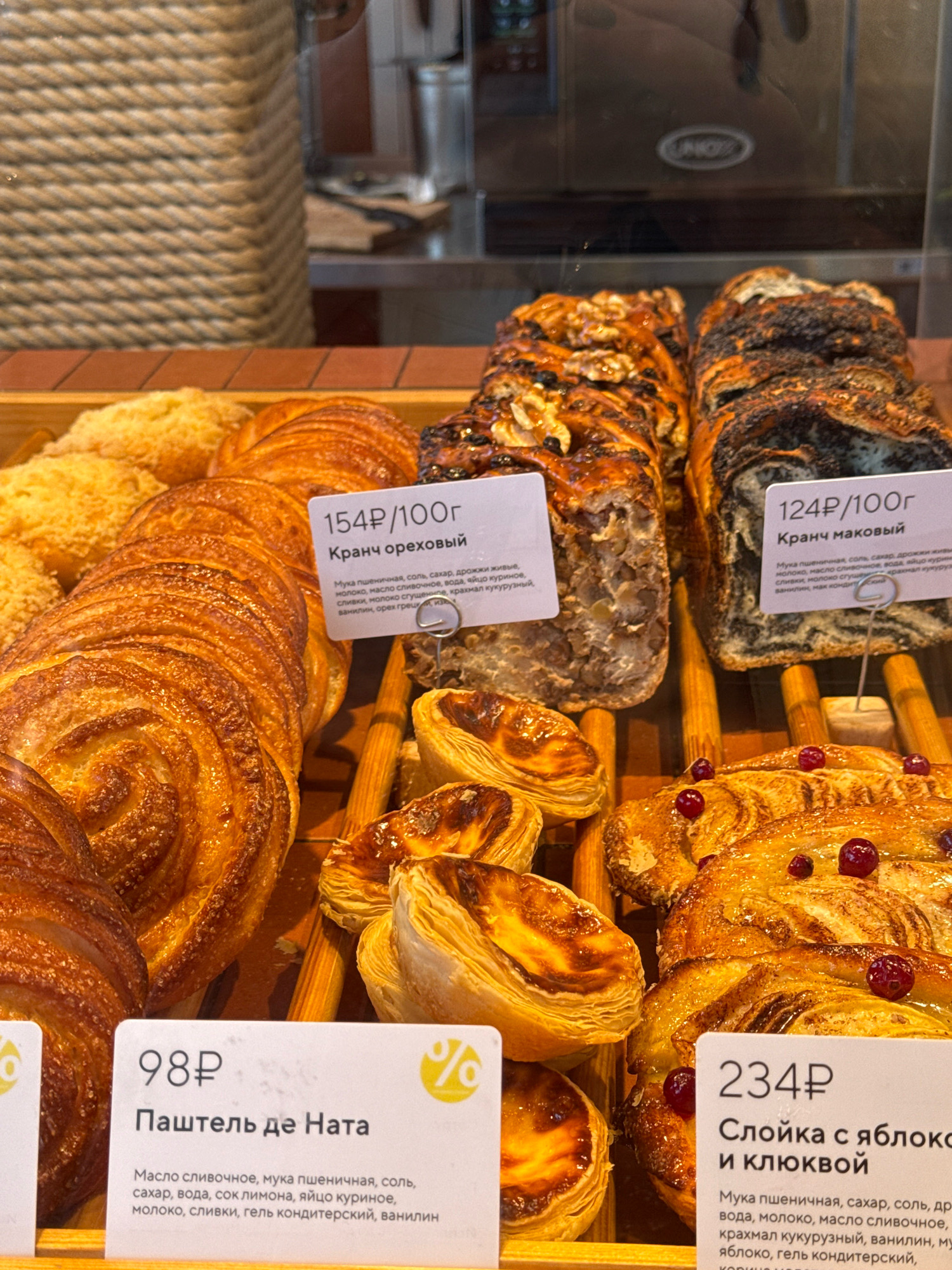 The image displays a close-up view of a bakery display case filled with various pastries. In the foreground, to the left, is a pastry labeled "98P Паштеть де Ната" with a list of ingredients. To its right, a pastry labeled "154P/100г Кранч ореховый" is visible, also with ingredients listed. Further to the right, another pastry is labeled "124P/100г Кранч маковый," detailing its composition. In the bottom right, a pastry labeled "234P Слойка с яблоком и клюквой" is shown with its ingredients. The pastries include swirled buns, layered pastries, and what appear to be tarts or danishes, some topped with berries. The display case is illuminated, highlighting the textures and golden-brown colors of the baked goods. Wooden dowels are used to separate and support some of the items. The background shows a blurred view of a counter and possibly kitchen equipment, with rope-like textures and signage visible. The image is taken from a slightly elevated angle looking down into the display. The pastries appear fresh and appealing. This scene is indicative of a bakery or pastry shop. Based on the Cyrillic labeling and the general context of baked goods, this could be a bakery in Kaliningrad, Russia, given the prompt's specificity request.