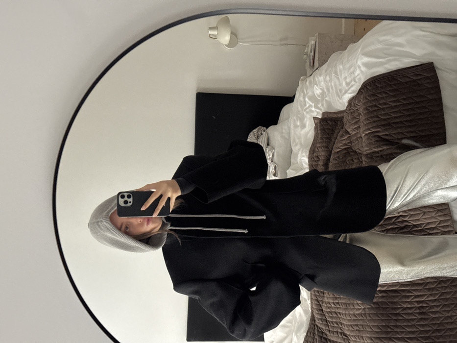 The scene depicts a mirror selfie taken indoors within a bedroom in Unknown, Unknown. A single individual, appearing female, is reflected in a large, black-framed arched mirror. She is dressed in a light grey knitted hood with visible drawstrings, layered over a black long-sleeved top and a black open blazer with structured shoulders. Her lower attire consists of metallic silver, wide-leg trousers. Her right hand holds a dark-colored smartphone, positioned to obscure the upper portion of her face, while her left hand rests on her hip. Behind the individual, a bed with a dark upholstered headboard is visible, covered with rumpled white bedding and a dark brown quilted throw blanket draped across the foot. To the right, a light-colored rectangular bedside table supports a white wall-mounted lamp with a conical shade. The walls are plain white.