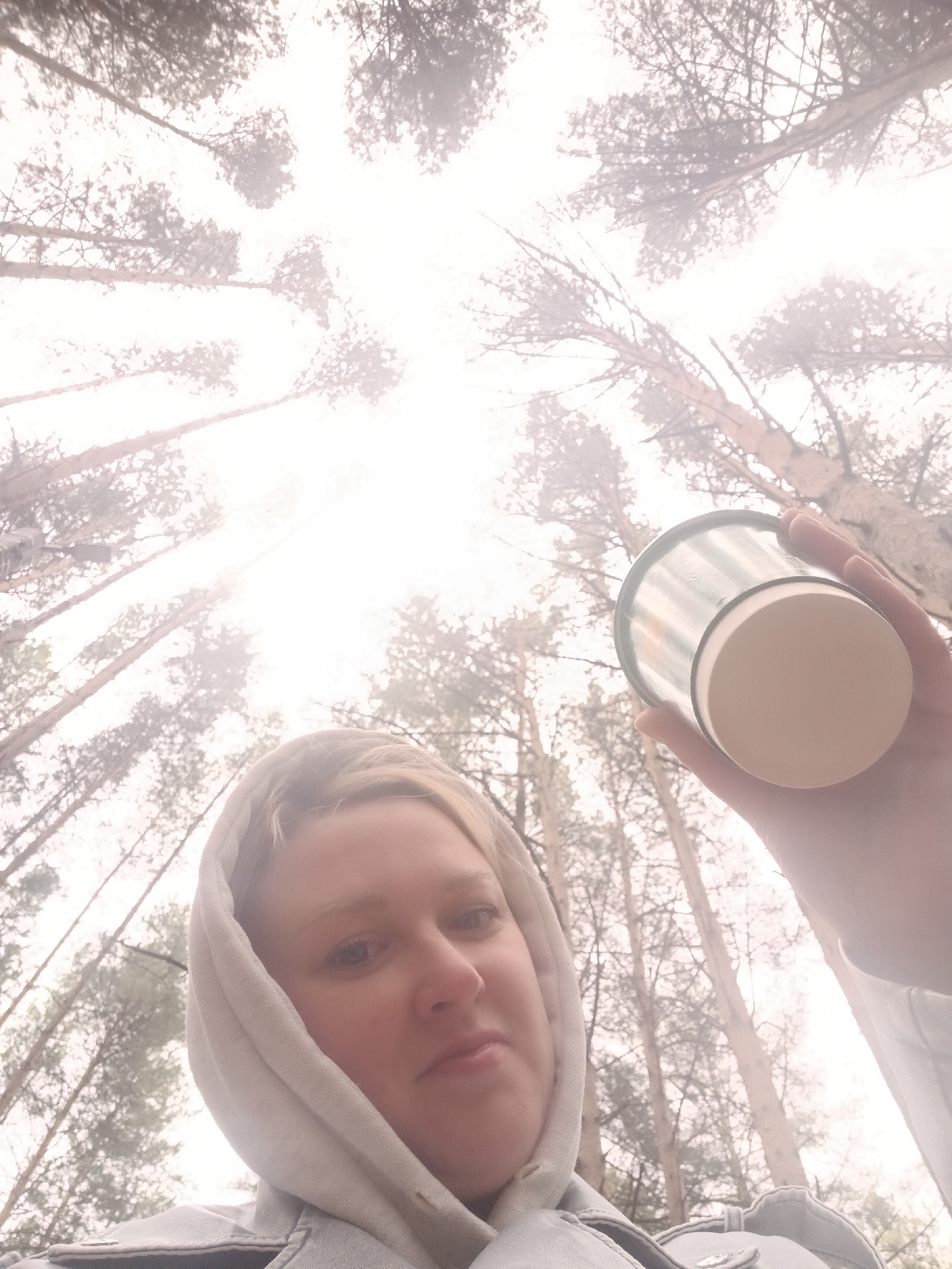 The image shows a woman in a light-colored hooded sweatshirt holding a cup up towards a forest canopy. The view is from below, looking up through tall, bare trees into a hazy, overcast sky. The location is the city of Chaikovsky, Russia. The woman is the primary subject, and the cup and forest are secondary elements. The interaction is the woman holding the cup while taking a selfie or photo. The environment is a misty forest.