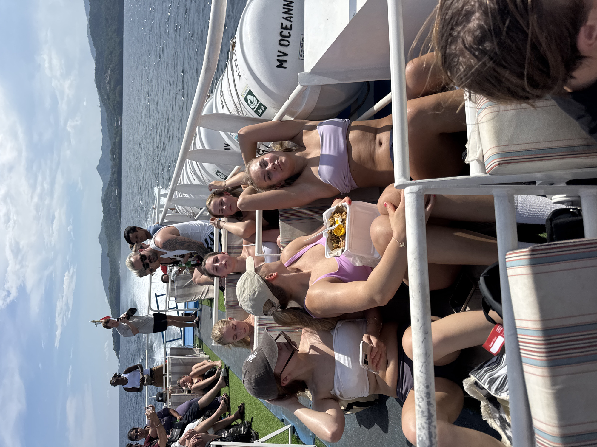 The image depicts a group of people on a boat, likely a ferry, sailing on a body of water with land visible in the distance. The setting is likely in the city of Unknown, Indonesia. The group consists of both men and women, some of whom are sunbathing or relaxing on lounge chairs. Several women are wearing bikinis. Some individuals are eating food from takeout containers. The boat appears to be moderately crowded. The environment is sunny and clear. There is a life raft labeled "MV OCEANNEX" on the boat.