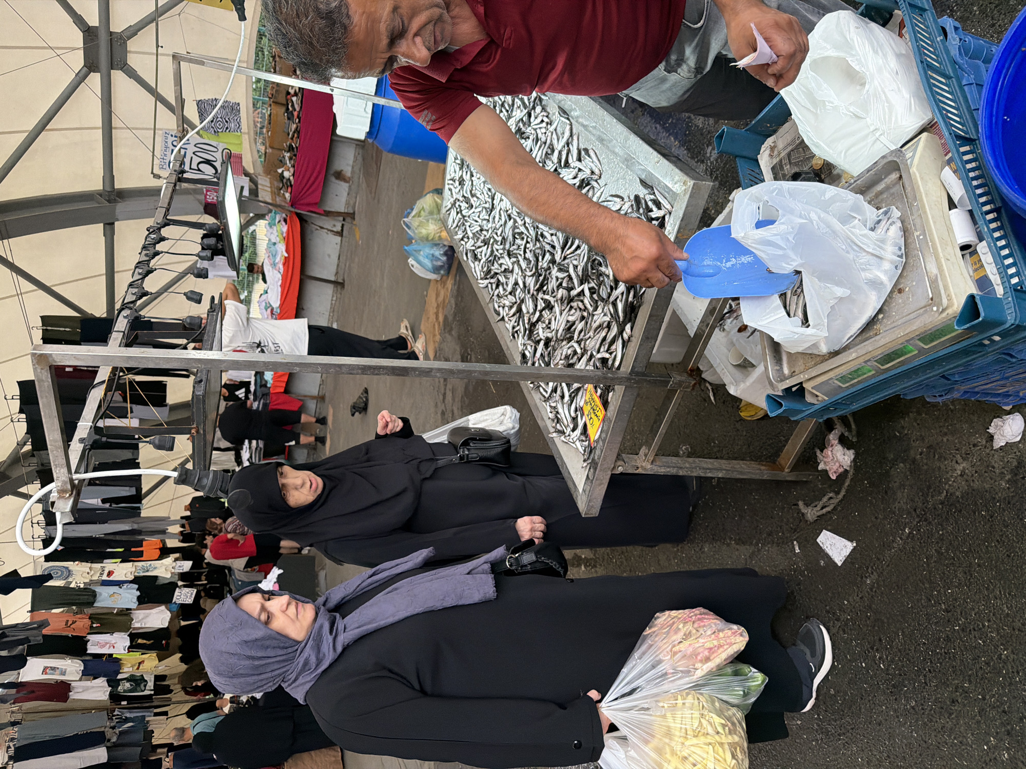 In Kadıköy, Turkey, a man in a red polo shirt and gray pants is selling small fish from a stainless steel table. He is using a blue plastic scoop to transfer fish into a plastic bag for a customer. Two women, dressed in black abayas and hijabs, are standing nearby. One woman is wearing a purple hijab and carrying a large plastic bag filled with vegetables. The other woman, wearing a black hijab, is looking at the fish. The market appears to be outdoors under a white tent structure. Clothes are hanging on racks in the background, and other market stalls are visible. The ground is wet and appears to be asphalt or concrete.