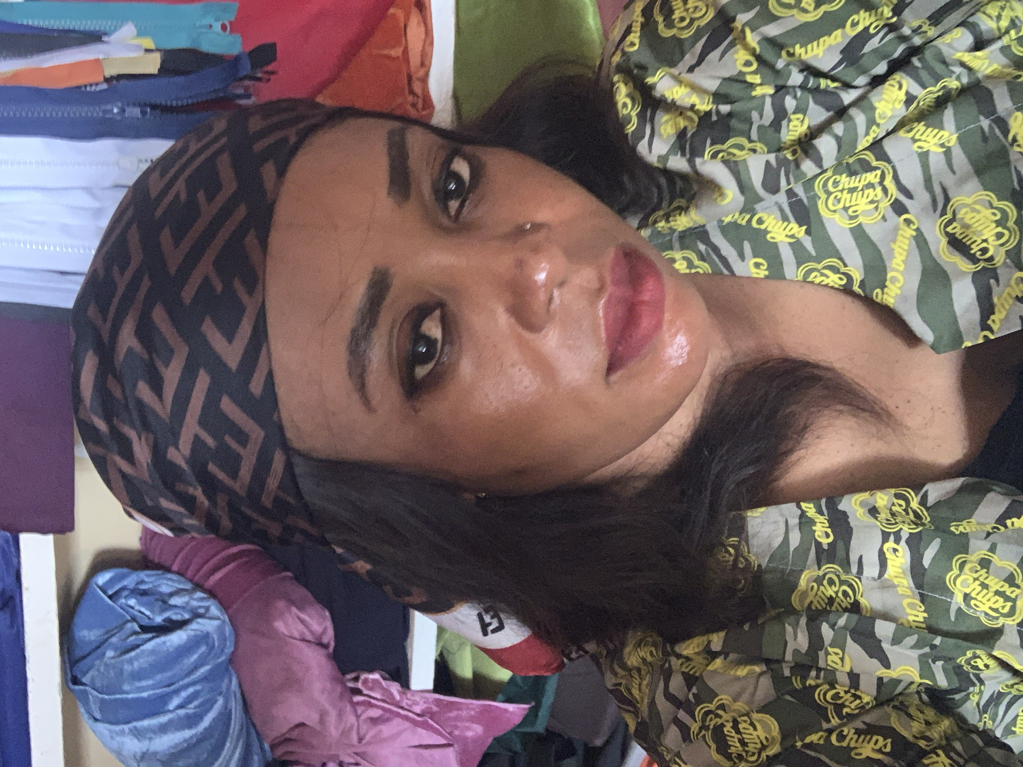An individual is captured in a close-up, tilted portrait, seemingly lying down and looking directly at the camera. The subject has dark skin, dark eyes, and dark hair, with defined eyebrows and reddish-pink lipstick. A small nose piercing is visible on their left nostril. They are wearing a dark brown head covering or scarf patterned with repeating, interlocking F-shaped logos in a lighter brown hue. Their attire includes a camouflage-patterned garment, likely a shirt, featuring multiple yellow "Chupa Chups" logos.
The background is a cluttered indoor space, characteristic of a textile workshop or retail area in Sabon Tasha, Nigeria. To the left of the subject's head, there's a stack of various colored zippers (including blue, white, orange, and light blue). Below these, several rolls or piles of fabric in colors such as deep purple and blue velvet are visible. Other fabrics in various hues, including red, green, and pink, fill the rest of the frame, suggesting an environment rich in textile materials and crafting supplies.
The visible text "Chupa Chups," "Chupa Chu," "Chipa," "Chup," "Chips," and "dm" can be seen on the camouflage fabric of the individual's shirt. The lighting appears to be ambient indoor light, and no specific time of day or weather conditions are discernible. The person's expression is neutral and direct, indicating a posed photograph.