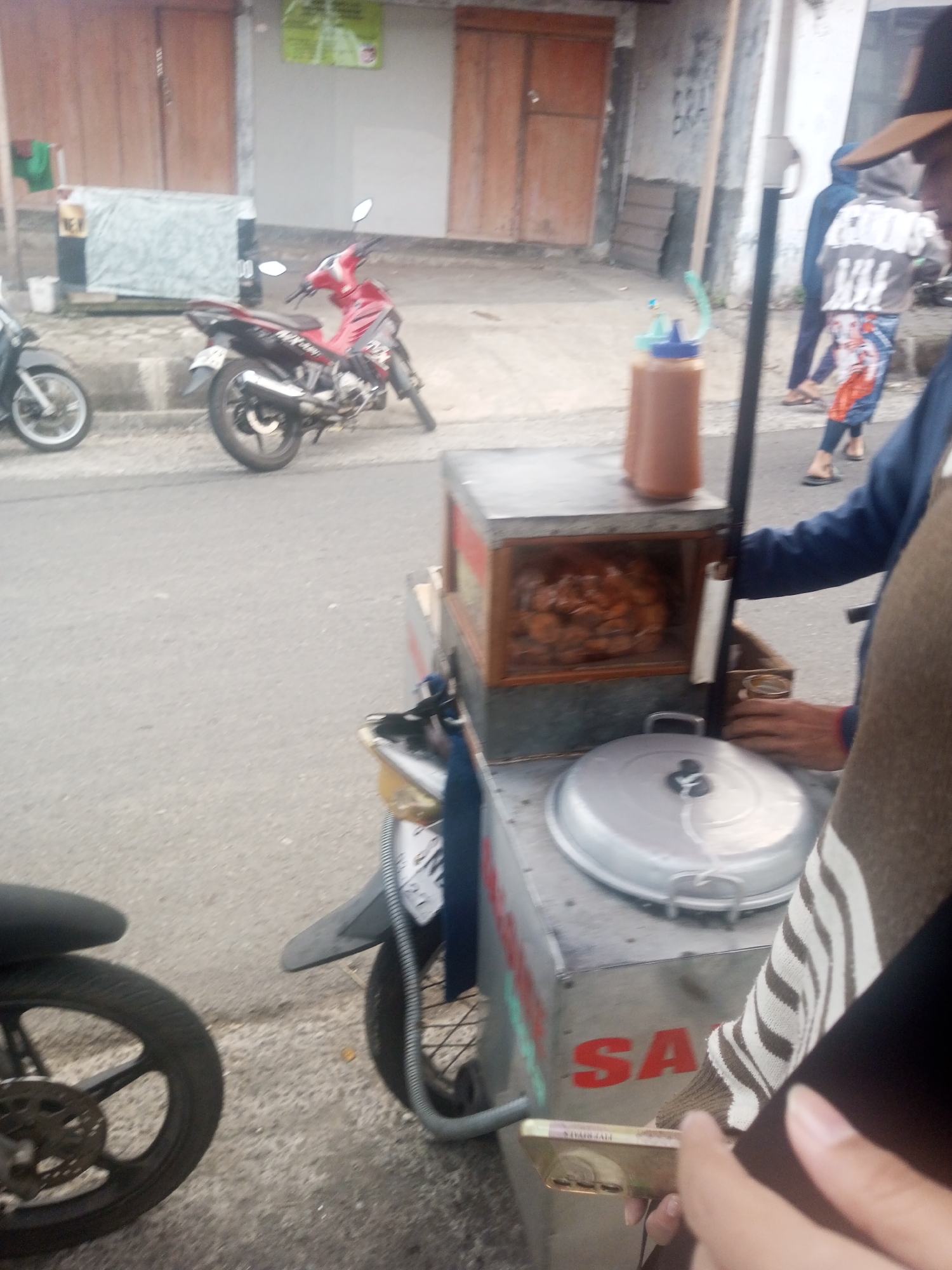 A food cart is on the side of a street in Kalibeber, Indonesia. The cart is stainless steel with a wooden-framed glass display case filled with food. On top of the cart is a large silver lid and a brown squeeze bottle. A person's arm is reaching towards the cart. In the background, a red motorcycle is parked on the street, and two people are walking in the distance. The street is paved, and buildings with wooden doors are visible on the left side of the image.