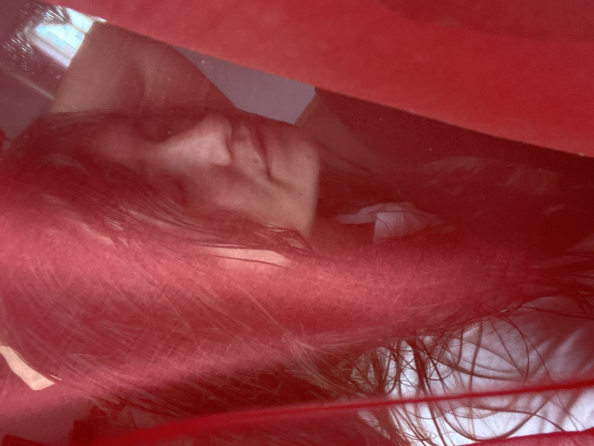 A close-up shot shows a person's face partially obscured by red fabric, possibly a veil or curtain. The person's eyes are closed, and their head is tilted slightly to the side, with one hand resting behind their head. The background is blurred, but appears to be a room with a window. The image was taken in the city of Perm, Russia.