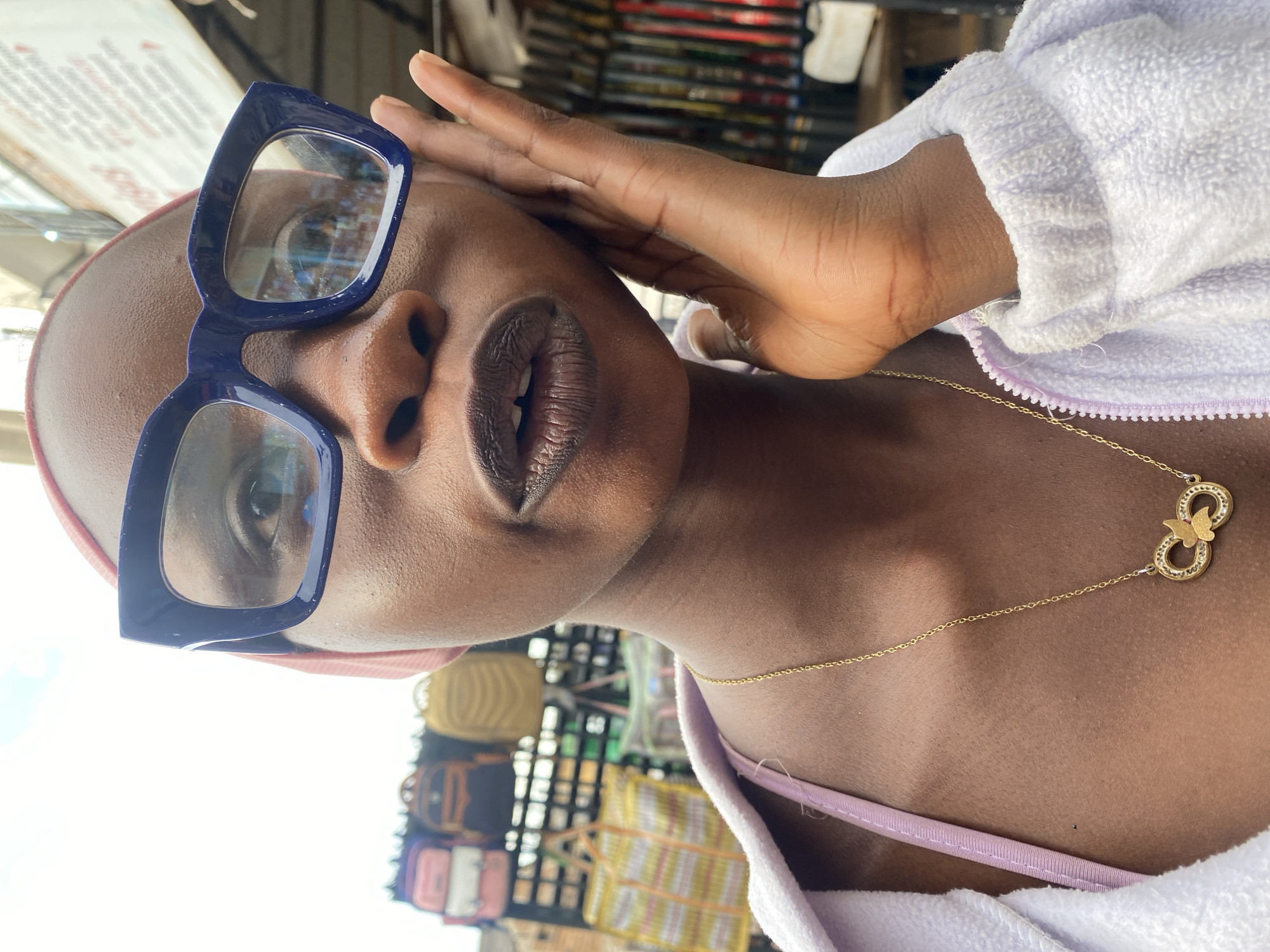 A close-up, waist-up outdoor shot shows a young Black woman from Jos, Nigeria, wearing large, dark blue, rectangular-framed eyeglasses and a pink headscarf. She has her right hand on her face, with her fingers spread, touching her cheek and temple. She wears a gold necklace with an infinity symbol pendant adorned with small, clear rhinestones and a golden butterfly. Her lips are slightly parted, and she appears to be looking towards the camera. She is also wearing a light purple zippered jacket over a pink bra or camisole. In the background, out of focus, are shelves stacked with various bags and other items, suggesting a market stall. The sky is visible above, with some clouds.