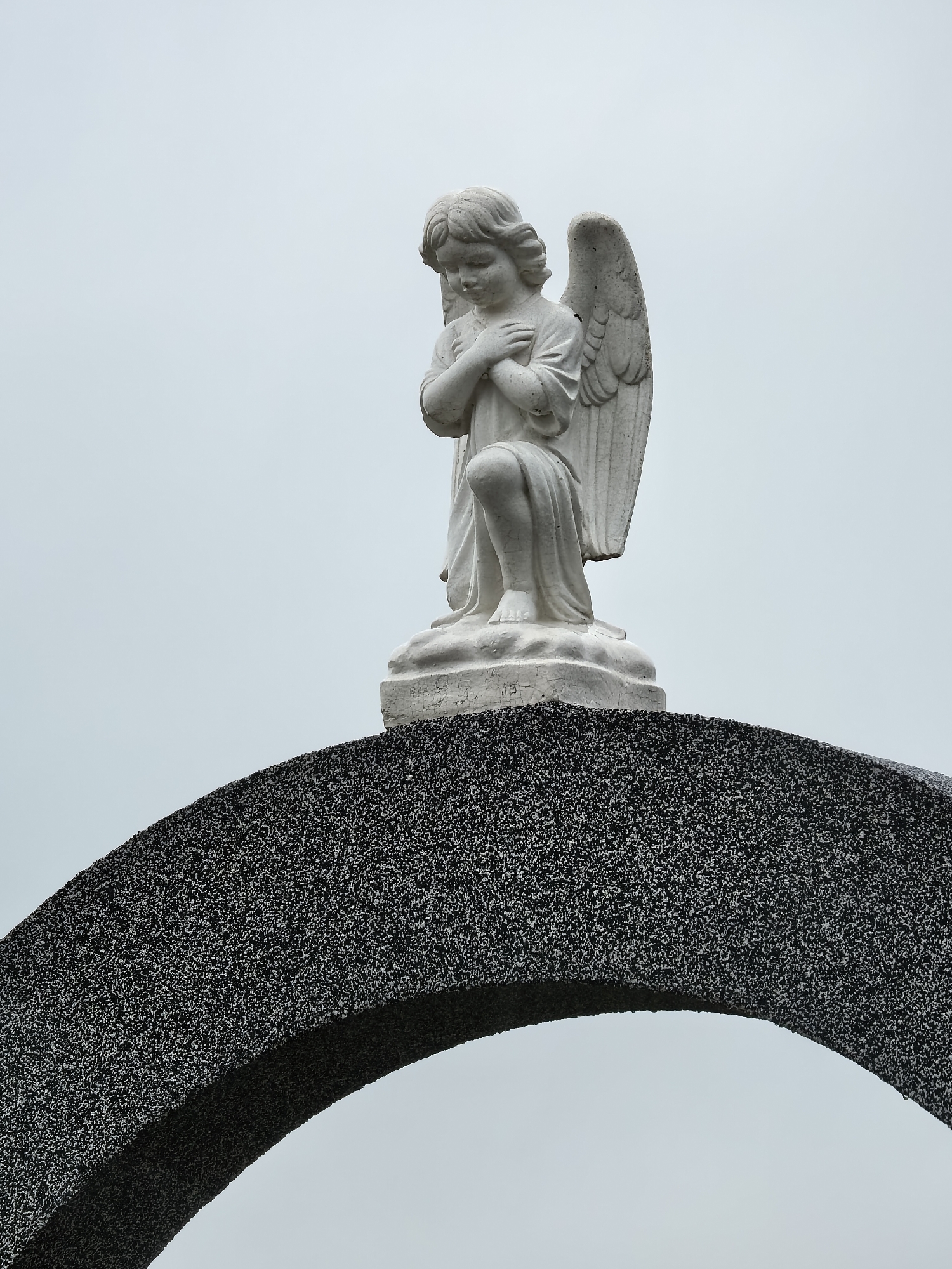 A white stone statue of an angel, appearing as a youthful child or cherub, is centered in the upper portion of the frame. The angel is depicted kneeling or crouching, with its head bowed and hands clasped together over its chest, suggesting a pose of prayer, contemplation, or mourning. Its wings are visible on its back, and its simple robe drapes around its legs. The statue rests on a white plinth, which shows some minor cracks or wear.
Below the statue is a large, dark, arch-shaped structure, made of a material with a rough, granular, speckled texture, appearing dark gray or black. The arch frames the lower part of the statue and extends outwards, creating a visual base for the figure. The sky behind the statue and within the arch is a uniform, light grey or pale blue, indicating an overcast day. There are no harsh shadows, suggesting soft, diffused light. The overall mood conveyed is one of solemnity, peace, or reverence. No specific activity or event is taking place beyond the static presence of the statue. No text is visible in the image.