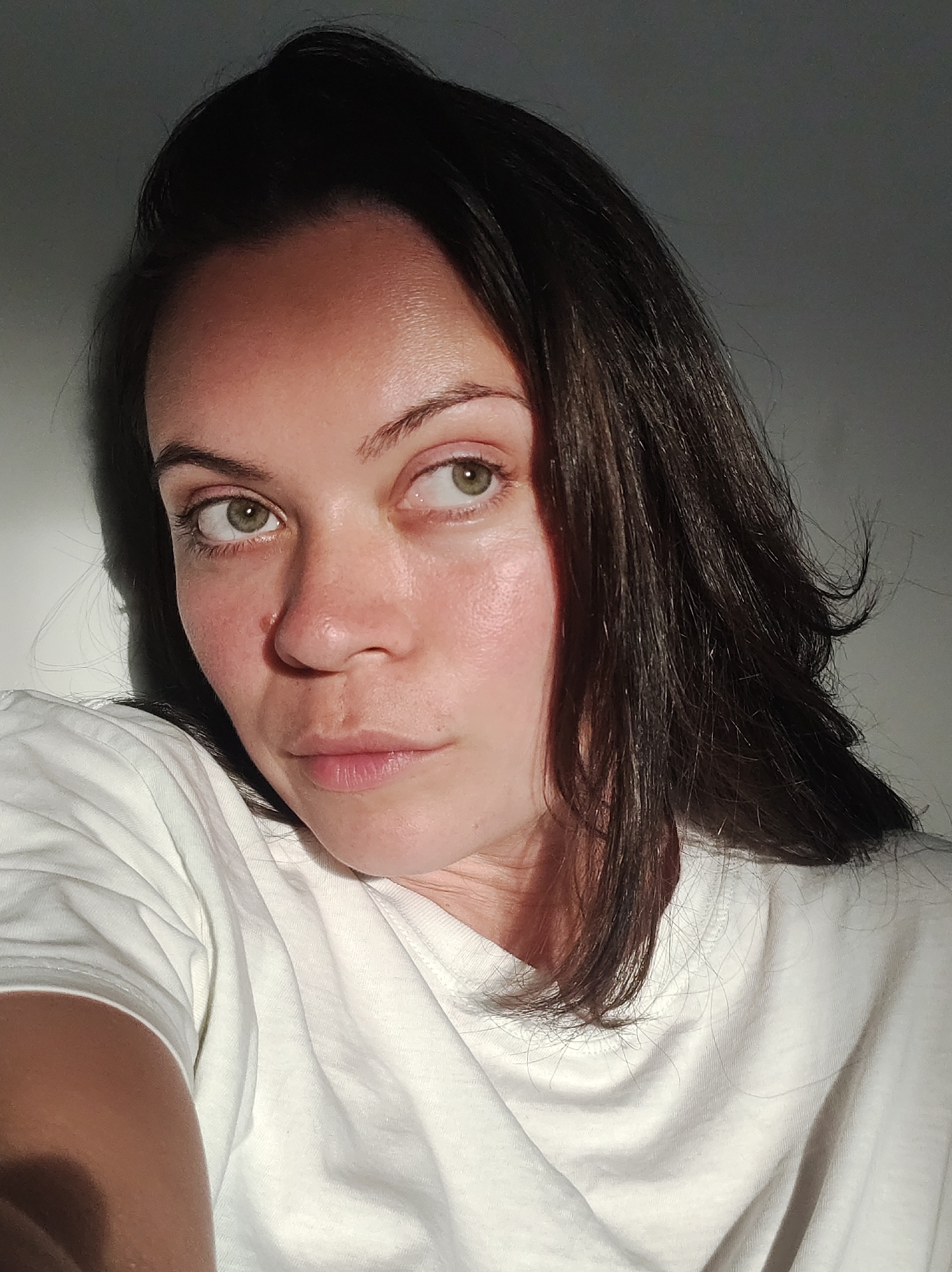 A close-up, high-angle, medium shot shows the face and upper body of a person with medium-toned skin and dark hair. The person is looking to their right with their head tilted slightly. They are wearing a white t-shirt, and the light in the scene is casting a shadow on the left side of their face and body. The background is a plain, gray wall.