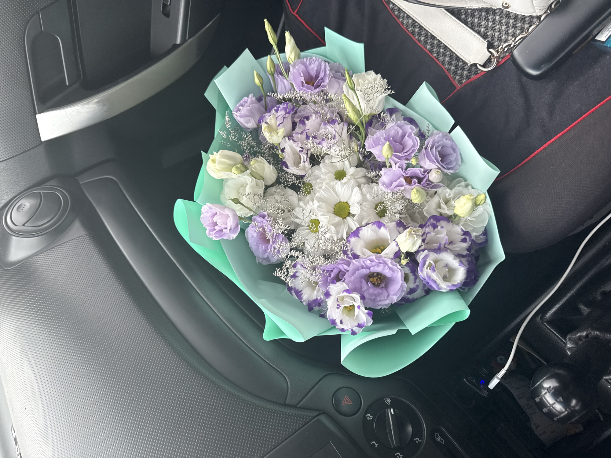 A bouquet of purple and white flowers is placed on the passenger seat of a car. The interior of the car is visible, with a dashboard and door panel. A charging cable is plugged into the car's power outlet, and a black gear shifter is also visible. The background suggests an indoor setting, possibly a garage or a parking lot, given the muted lighting and the car's interior. The image does not provide any specific indicators for location in Abaclia, Moldova.