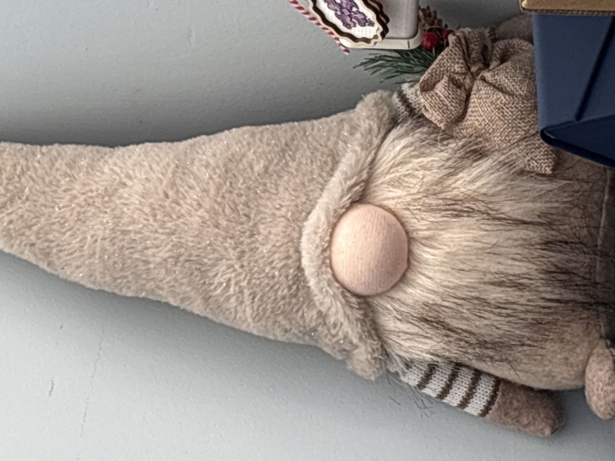 A close-up, horizontally oriented shot features a decorative plush gnome-like figure resting against a plain, light blue or grey wall in Sofia, Bulgaria. The gnome's prominent feature is a large, rounded, light pink fabric nose, positioned centrally in the image.
Above the nose, a soft, plush hat extends to the left, covered in light brown or tan fabric with subtle silvery flecks, suggesting a sparkly or glittery finish. Below the nose, a voluminous beard of long, greyish-brown faux fur cascades downwards and to the right. Part of a striped fabric element, possibly a sleeve or arm with horizontal brown and white stripes, is visible beneath the beard. A small, rounded piece of brown felt-like fabric is also seen near the bottom right, resembling a hand or foot.
In the upper right portion of the image, partially obscured, are festive decorative elements including small red berries and green pine needles. Adjacent to these, a crumpled piece of tan, burlap-textured fabric forms a decorative bow or embellishment. A dark blue, rigid, box-like object is visible on the far right edge. Above the gnome's hat, in the top left corner, a small section of a white object with a red and white striped ribbon is visible, hinting at further seasonal decor. The background wall is smooth and unadorned, providing a neutral backdrop. No text is visible.