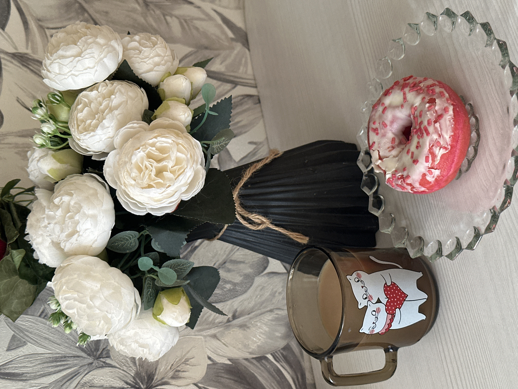 A still life photograph depicts a bouquet of white roses tied with twine resting on a surface with a floral pattern. Next to the bouquet is a dark-tinted mug with a cartoon-like image of two cats wearing red-spotted apparel. To the right of the mug and bouquet, there is a donut with pink frosting and white sprinkles on a glass plate. The location is in Chaikovsky, Russia.