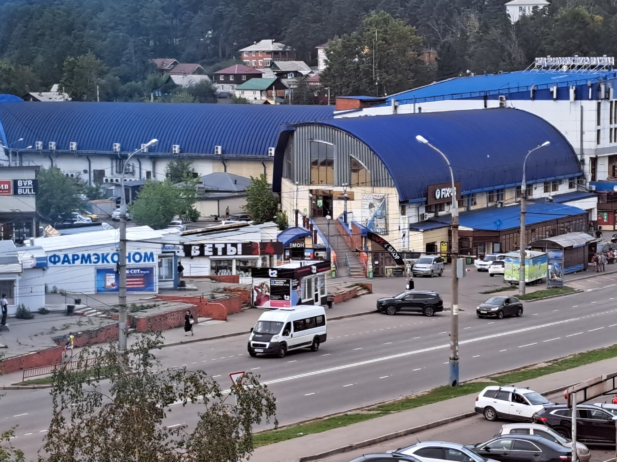 The image shows a street scene in Irkutsk, Russia, on a cloudy day. A large blue building with a curved roof dominates the background. In front of it, there are several smaller buildings and shops, including a pharmacy with a blue sign that reads "ФАРМЭКОНОМ" (PharmEconom). A white van is driving on the street, and several cars are parked or moving. People are visible walking on the sidewalk and near the shops. The street is paved, and there are sidewalks and traffic lights. Trees and foliage are present on the sides of the street and in the background, indicating a somewhat green urban environment. The overall impression is that of a commercial area with some residential buildings in the distance, nestled against a backdrop of a wooded hill.