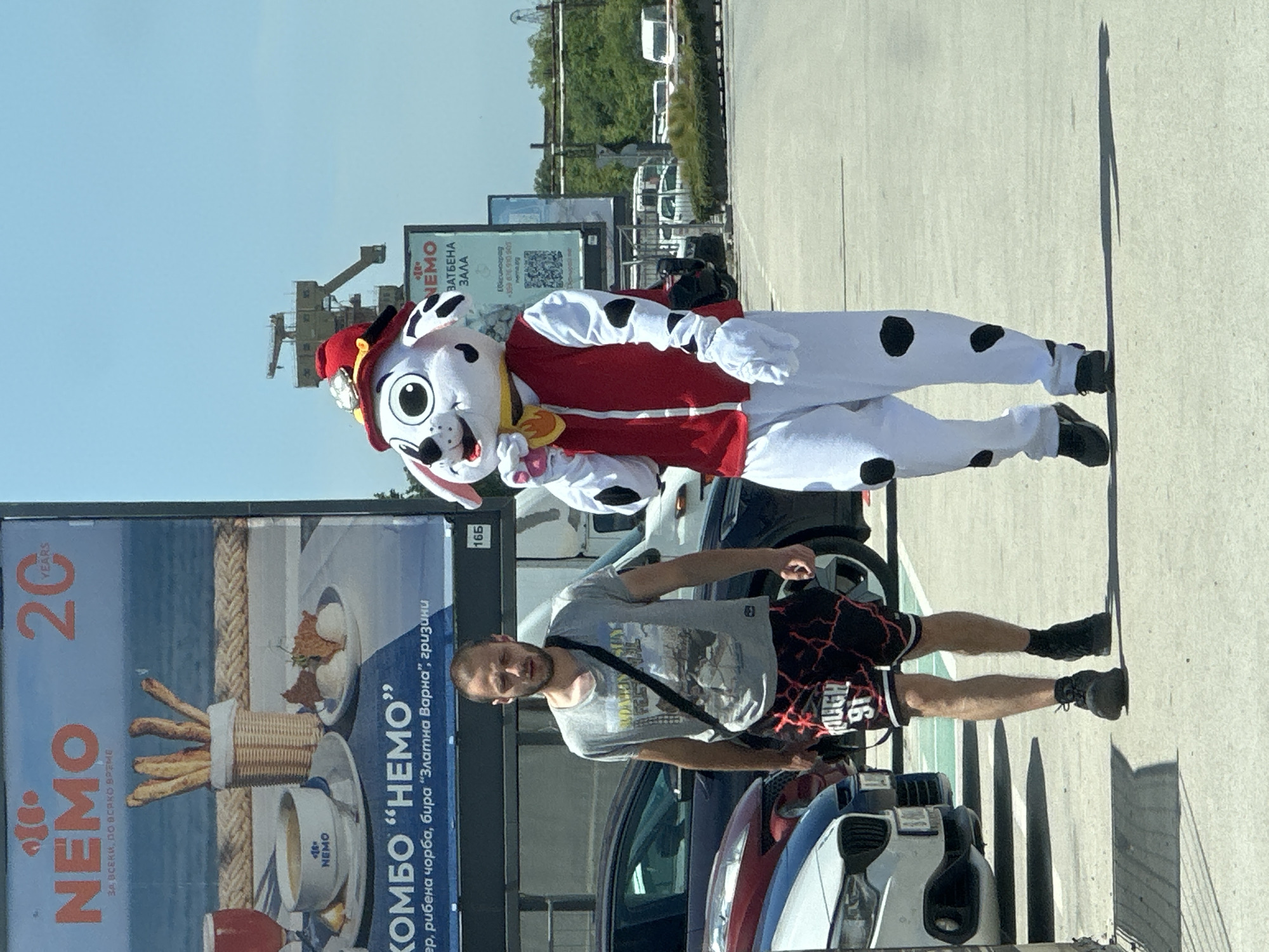The image shows a man standing next to a Paw Patrol character costume in what appears to be an outdoor parking area. The Paw Patrol character is likely Marshall, given the red firefighter hat and spotted Dalmatian design of the suit. The man is wearing a gray T-shirt, black and red shorts, and black shoes. A black strap of some kind is across his chest.
Behind the man and the costume, there are several cars and two large advertising billboards. One billboard advertises "NEMO", a restaurant or event location, with the tagline "20 years" and the description of a combo meal including fish soup and beer. The other sign also appears to advertise "NEMO", with the text "Ватбена зала," likely a venue for weddings. The phrase "Златна Варна" (Golden Varna) appears on one of the billboards, indicating that this is likely in or near the city of Varna, Bulgaria.