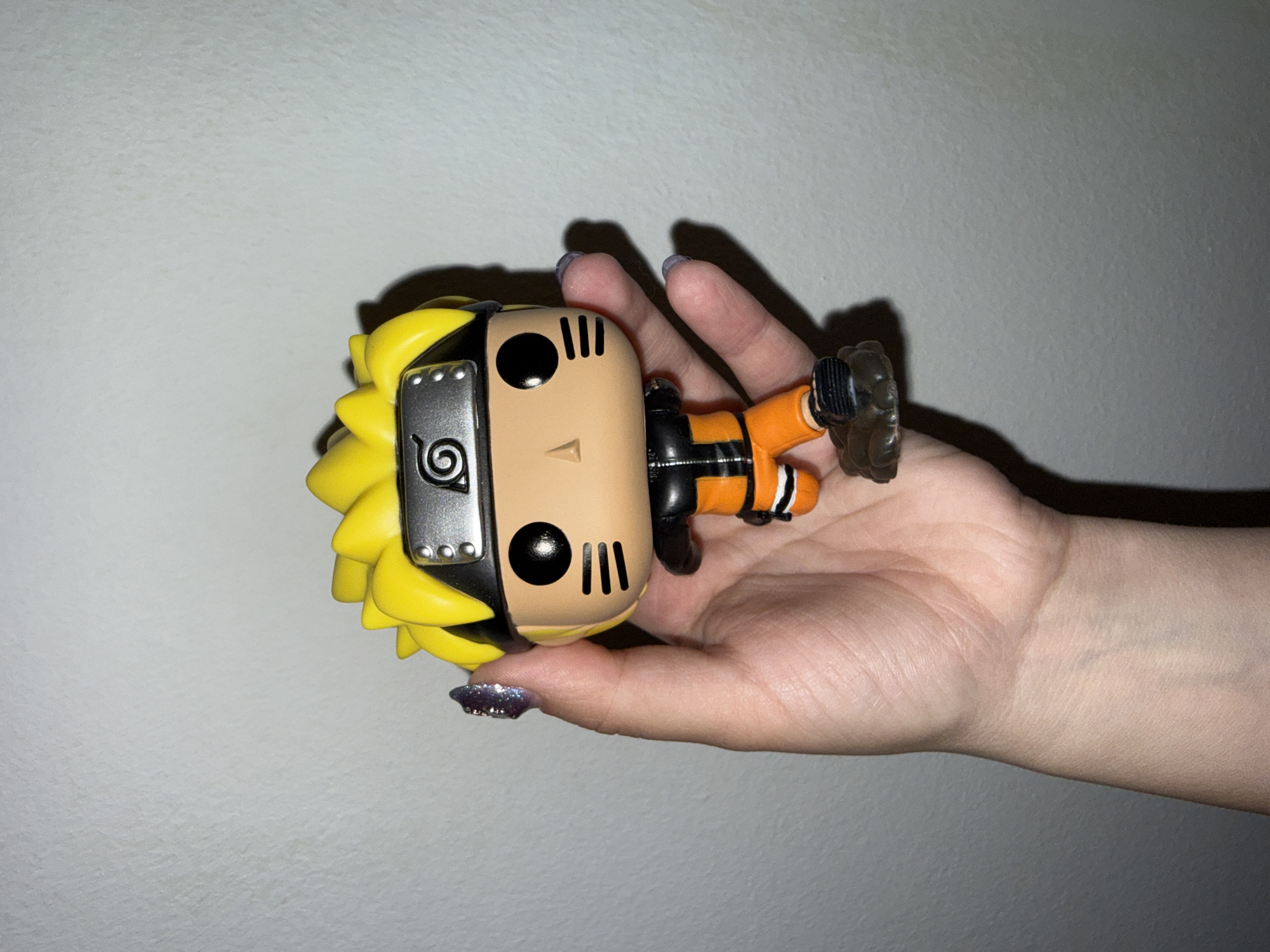The image displays a human hand holding a Funko Pop figure of Naruto Uzumaki against a plain, light-colored wall. The hand holds the figure horizontally, with the character appearing to lie on its back.
The Funko Pop figure is clearly identifiable as Naruto Uzumaki from the anime and manga series *Naruto*. It features the character's iconic bright yellow spiky hair, a black headband with a metallic plate engraved with the Konoha village symbol (a spiral), orange jumpsuit clothing, and distinct whisker marks on its cheeks. The figure has the typical large, black, circular eyes characteristic of Funko Pop designs.
The hand holding the figure is light-skinned, with visible knuckles and skin texture. The fingernails are short and well-kept, with some adorned with a shimmering purple glitter polish. Shadows from both the hand and the figure are cast onto the wall, indicating a light source illuminating the scene from the front or above. The background wall is a uniform light grey or off-white color, showing a slight, subtle texture.
The setting appears to be indoors, likely within a residence or similar environment, situated in the city of Zagreb, Croatia. No specific time of day or weather conditions are discernible from the image, and there is no visible text other than the character's design elements. The image focuses on the collectible item being casually presented or held.