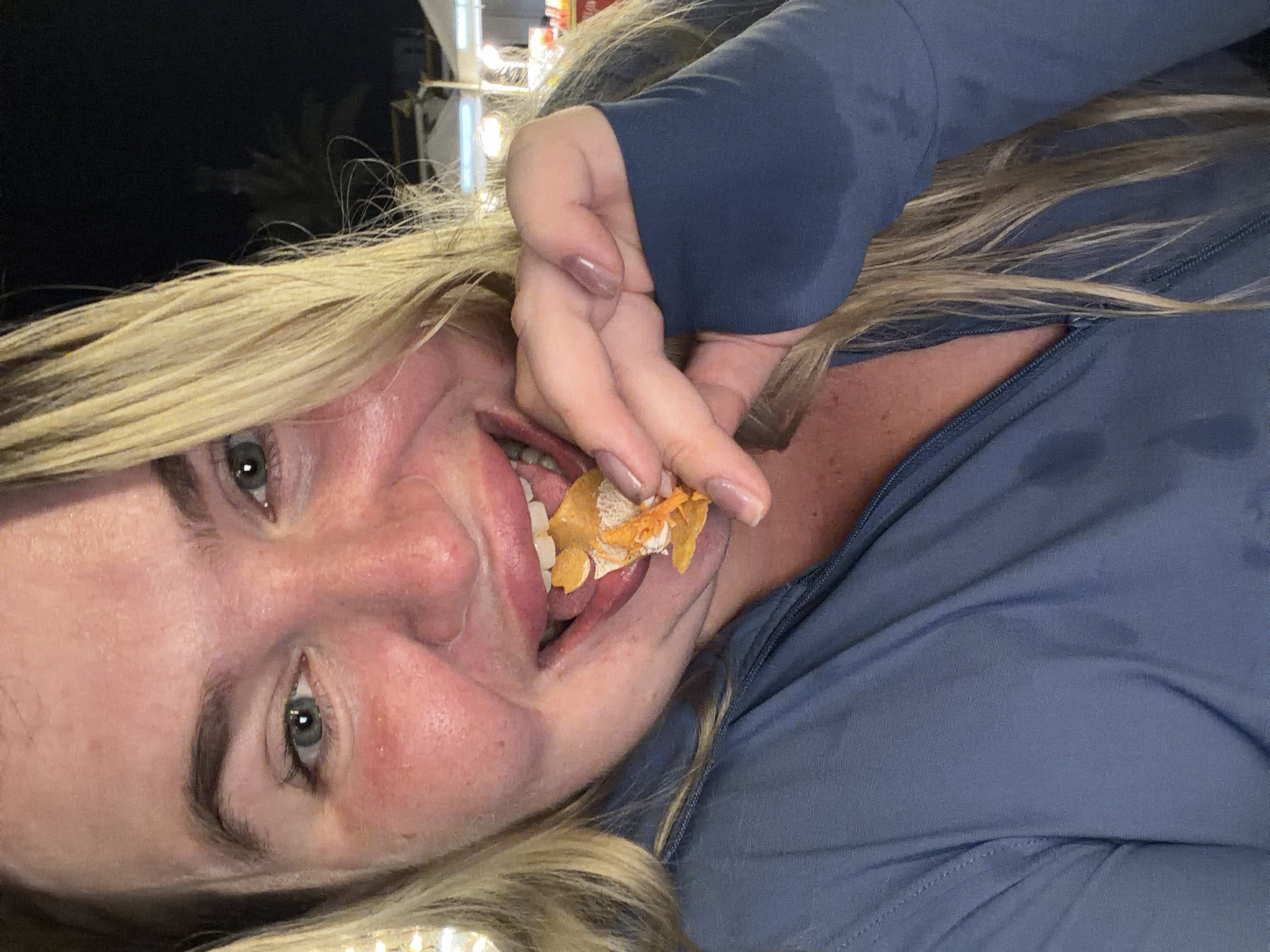 In this close-up, selfie-style image, a woman with blonde hair and light-colored eyes is shown from the chest up, eating. She is wearing a blue zippered jacket. Her mouth is open, and she is holding a piece of food, which appears to be a fritter or a similar fried item, to her lips with her right hand. Her fingernails are light-colored and appear to be painted. The background is dark, suggesting it is nighttime, with blurred lights and what appear to be stalls or tents, possibly indicative of a night market. The woman's skin is flushed, particularly on her cheeks. The scene has the appearance of being outdoors, possibly in a public area within Phuket City Municipality, Thailand, given the context.