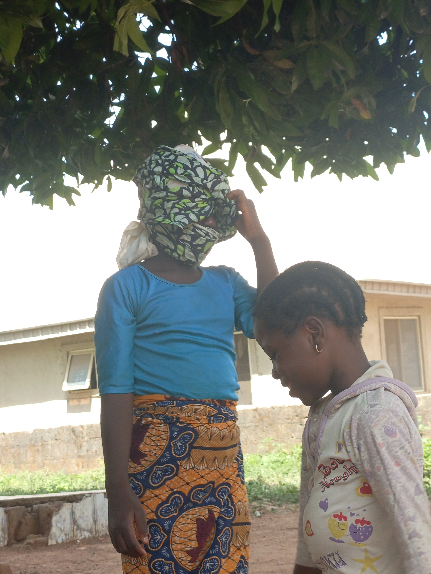 Two girls are standing outdoors in Akwanga, Nigeria. The taller girl wears a blue shirt, an orange and blue patterned wrap skirt, and a green and black head wrap obscuring her face. She has her right hand raised to her head. The shorter girl stands to the right and is facing right. She wears a light colored hooded sweatshirt. The background includes a building, some grass, and a tree with green leaves overhanging the scene. The ground appears to be a dirt path.