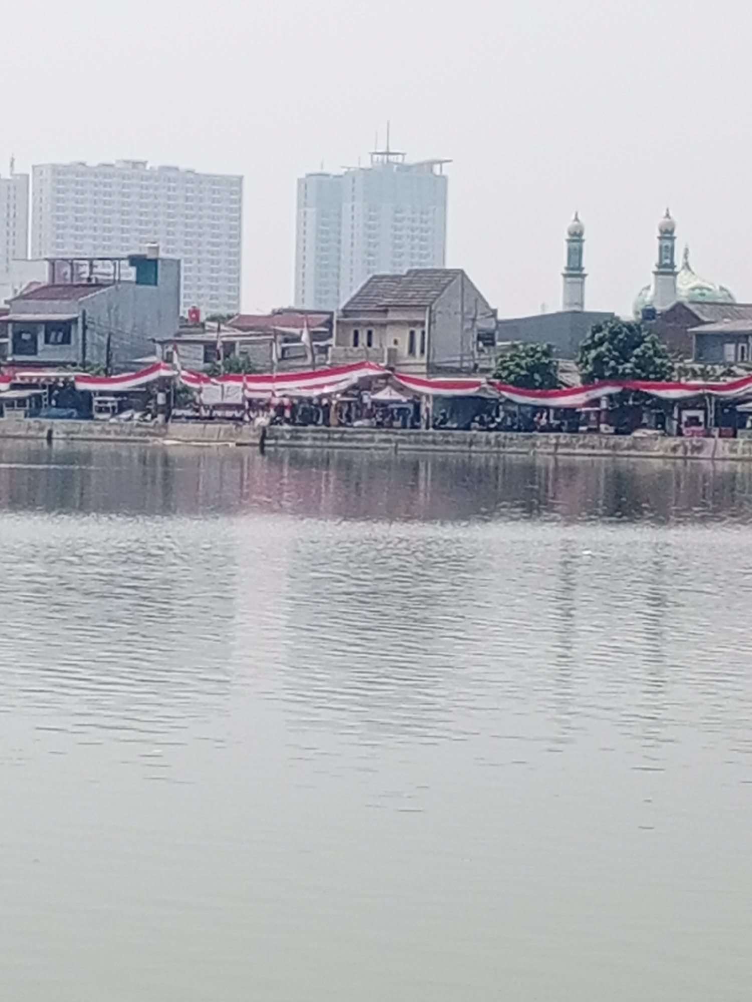 The image shows a waterfront scene in Depok, Indonesia, featuring a river in the foreground with ripples on its surface. Along the bank, a row of buildings with red and white awnings is visible, some of which appear to be small shops or stalls. Red and white banners are draped across these structures, suggesting a festive or patriotic occasion. In the background, several tall apartment buildings and a mosque with green domes and minarets rise against a hazy sky. The overall impression is of an urban environment with a mix of residential, commercial, and religious architecture.
