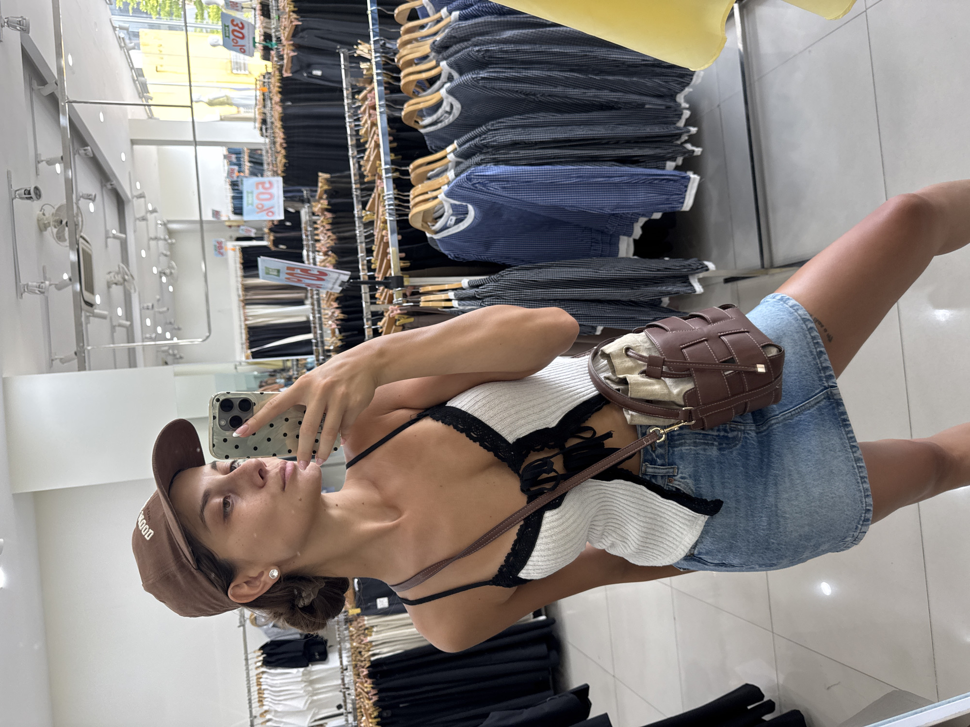 A woman stands in a clothing store, likely in Đà Nẵng, Vietnam. She is taking a selfie in front of a mirrored wall. She is wearing a brown cap, a white and black top, and a denim skirt. She has a small brown purse slung over her shoulder. The store has rows of clothing racks filled with shirts and other garments. Signs indicating price reductions, such as "20%" and "30%," are visible on the racks. The floor is tiled.